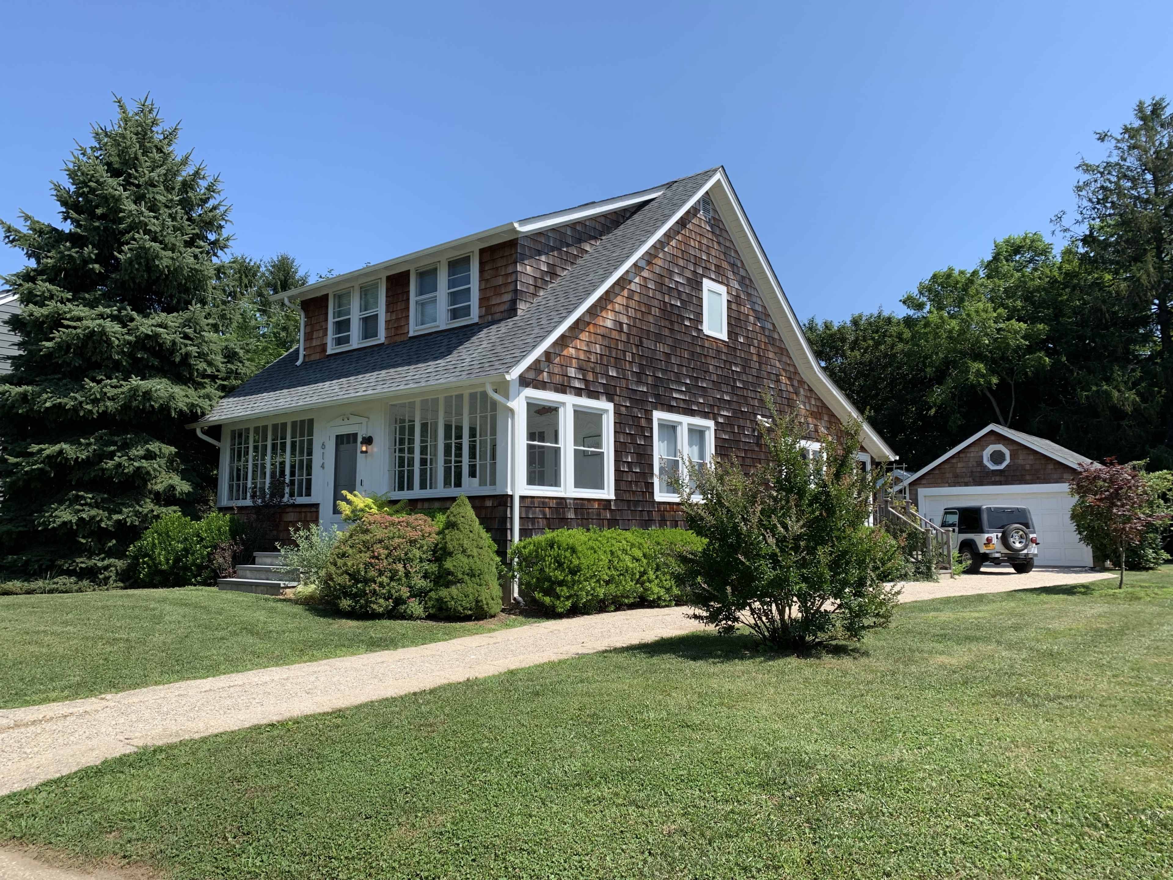 A two-story shingle-style house with a front porch is surrounded by a manicured lawn and shrubs, with a detached garage in the background.