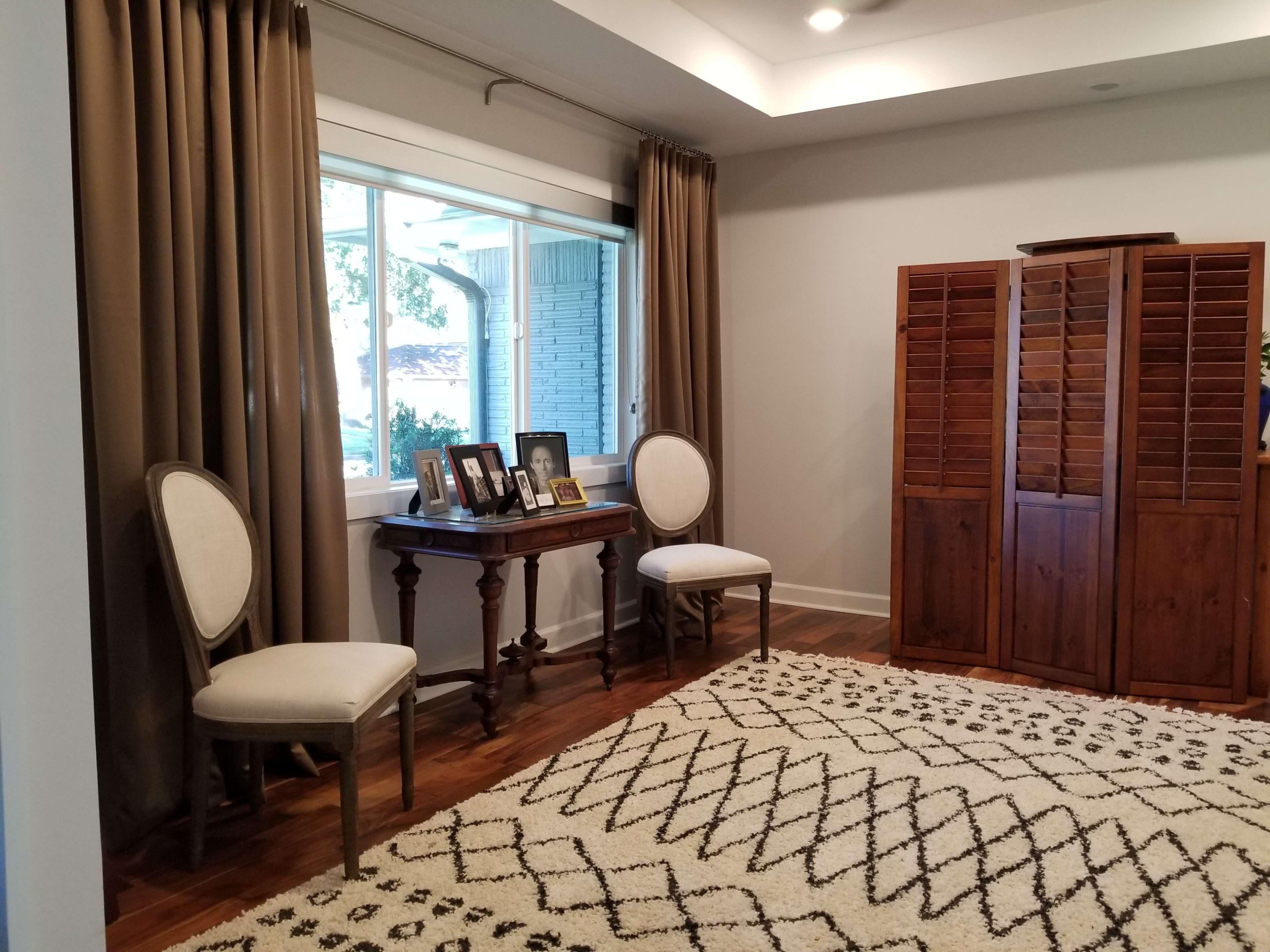 A room with two upholstered chairs, a small wooden table displaying framed photos, and a wooden cabinet next to a large window.