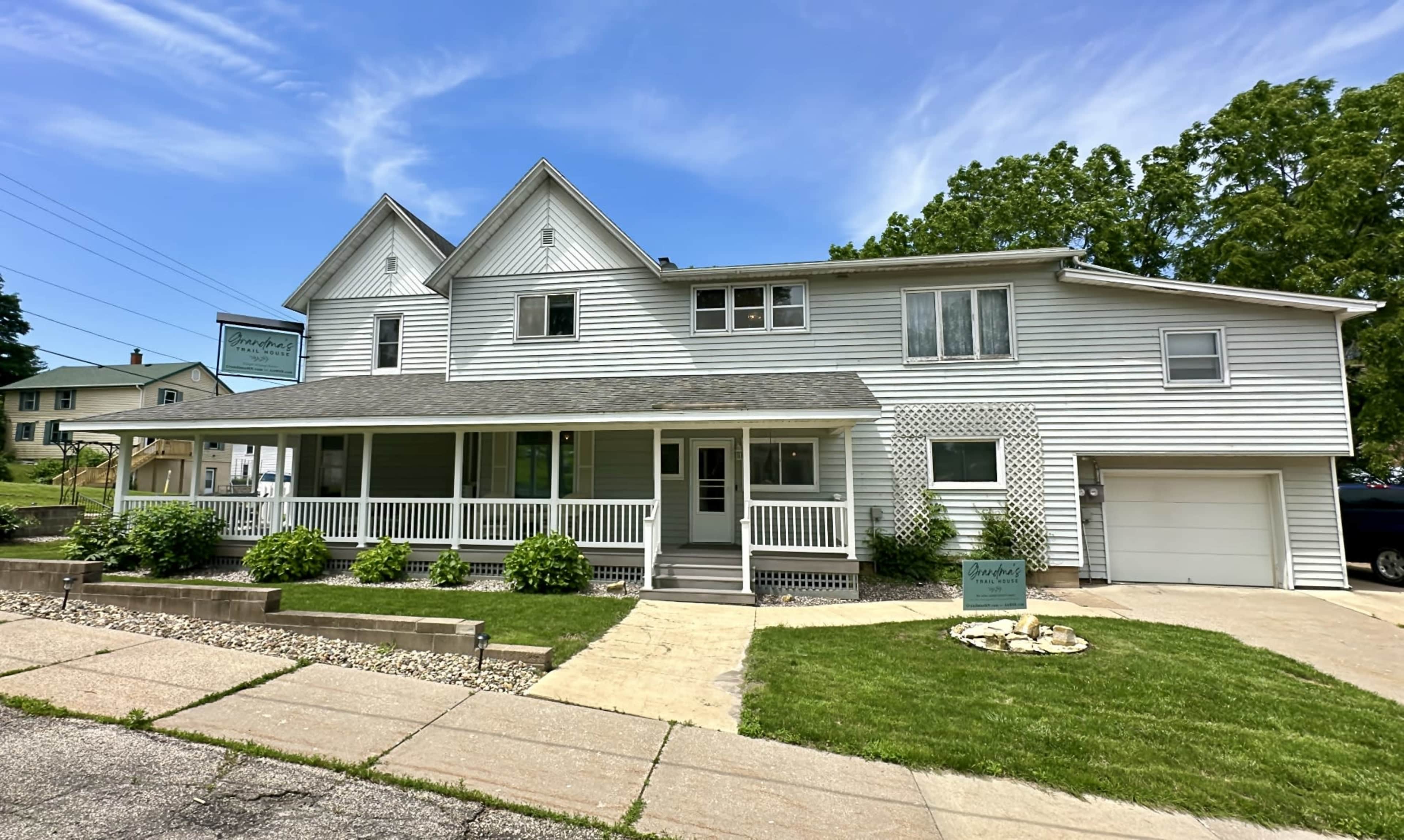 A light-colored, two-story house with multiple gables features a front porch and a sign outside, surrounded by a well-maintained lawn and sidewalk.