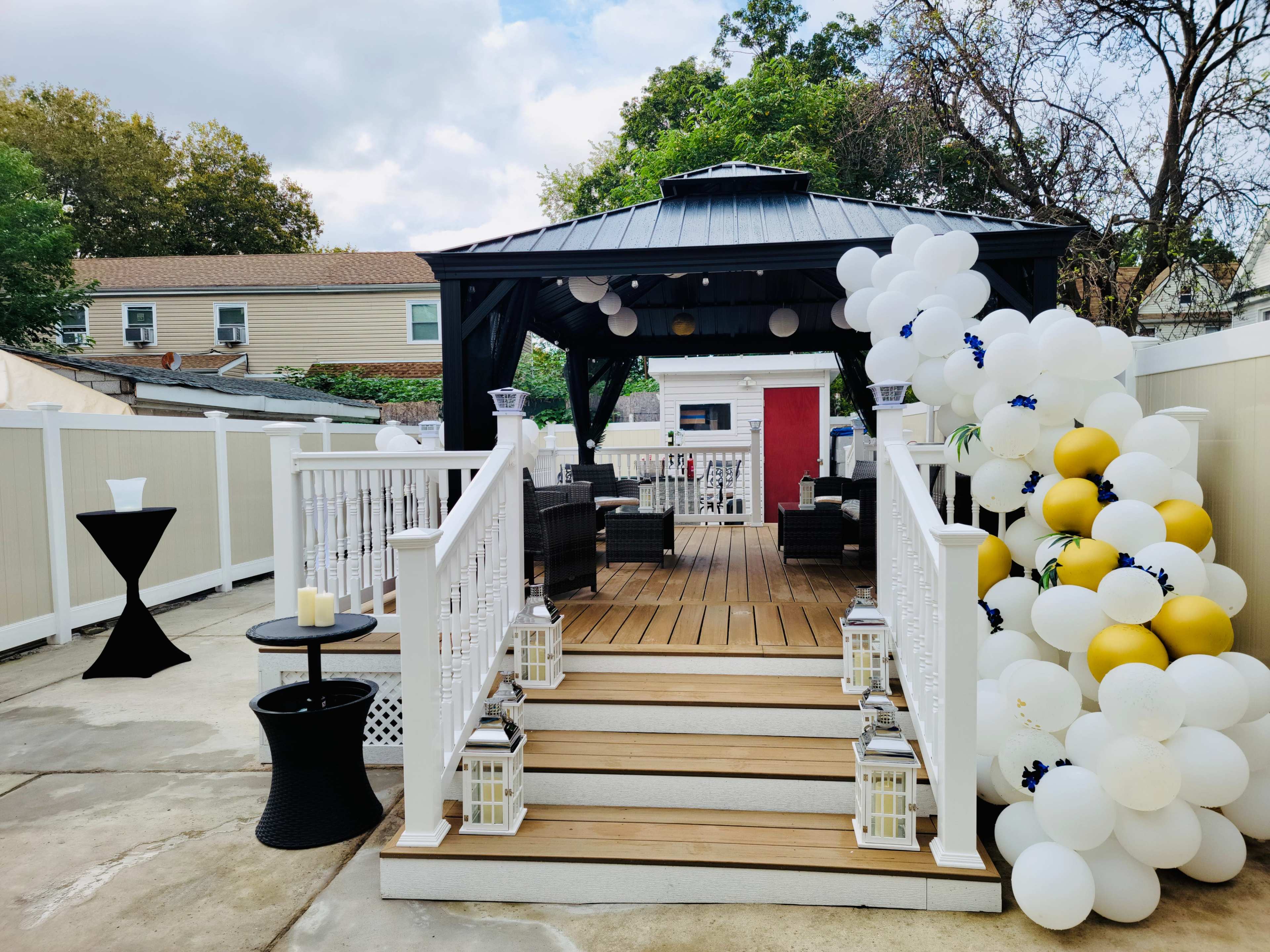 The image shows a wooden deck leading to a gazebo, decorated with balloons and featuring lounge seating, surrounded by a white fence.