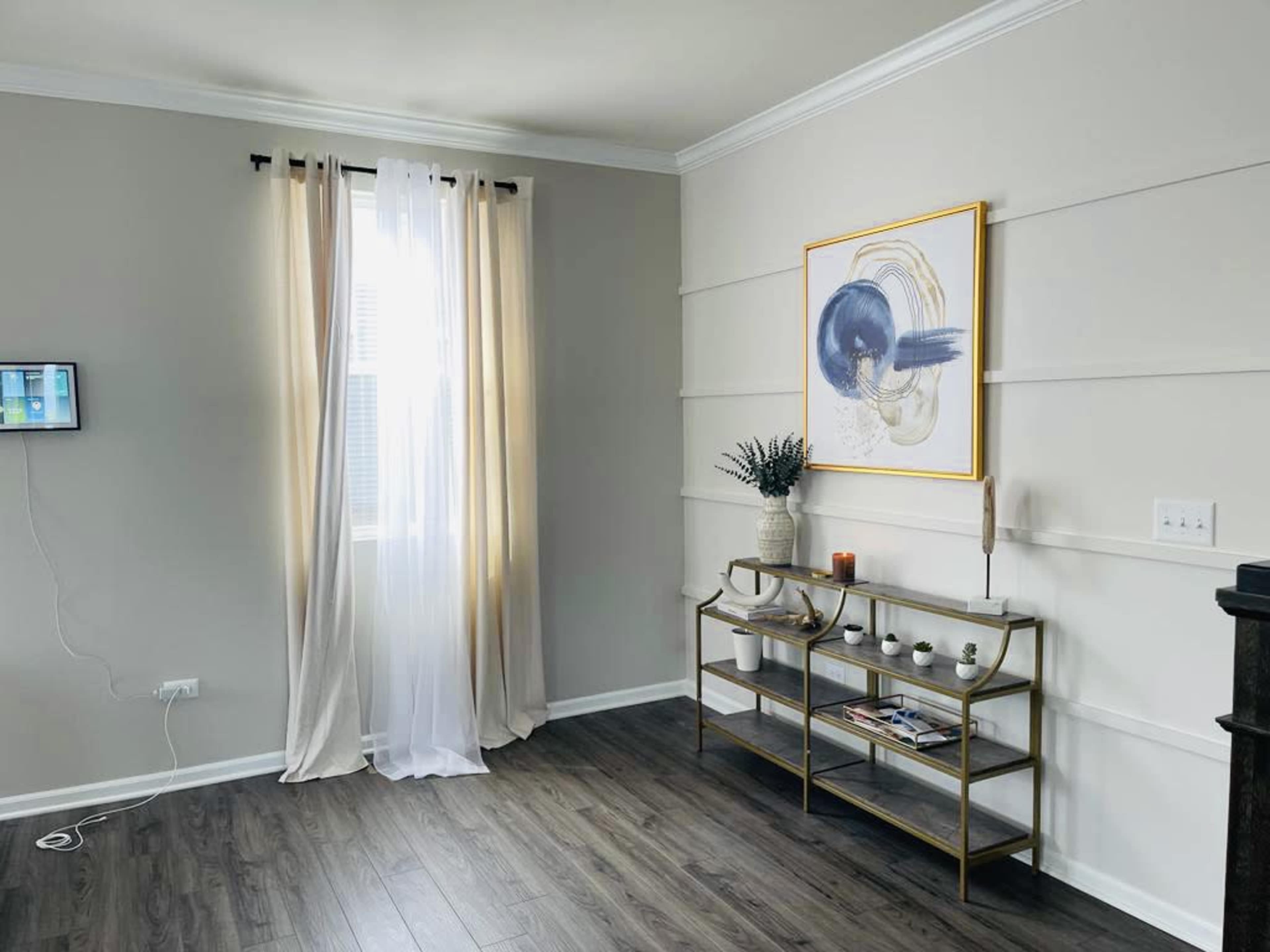 A room with light-colored walls features a large window with sheer curtains, a framed artwork on the wall, and a glass console table displaying decorative items.