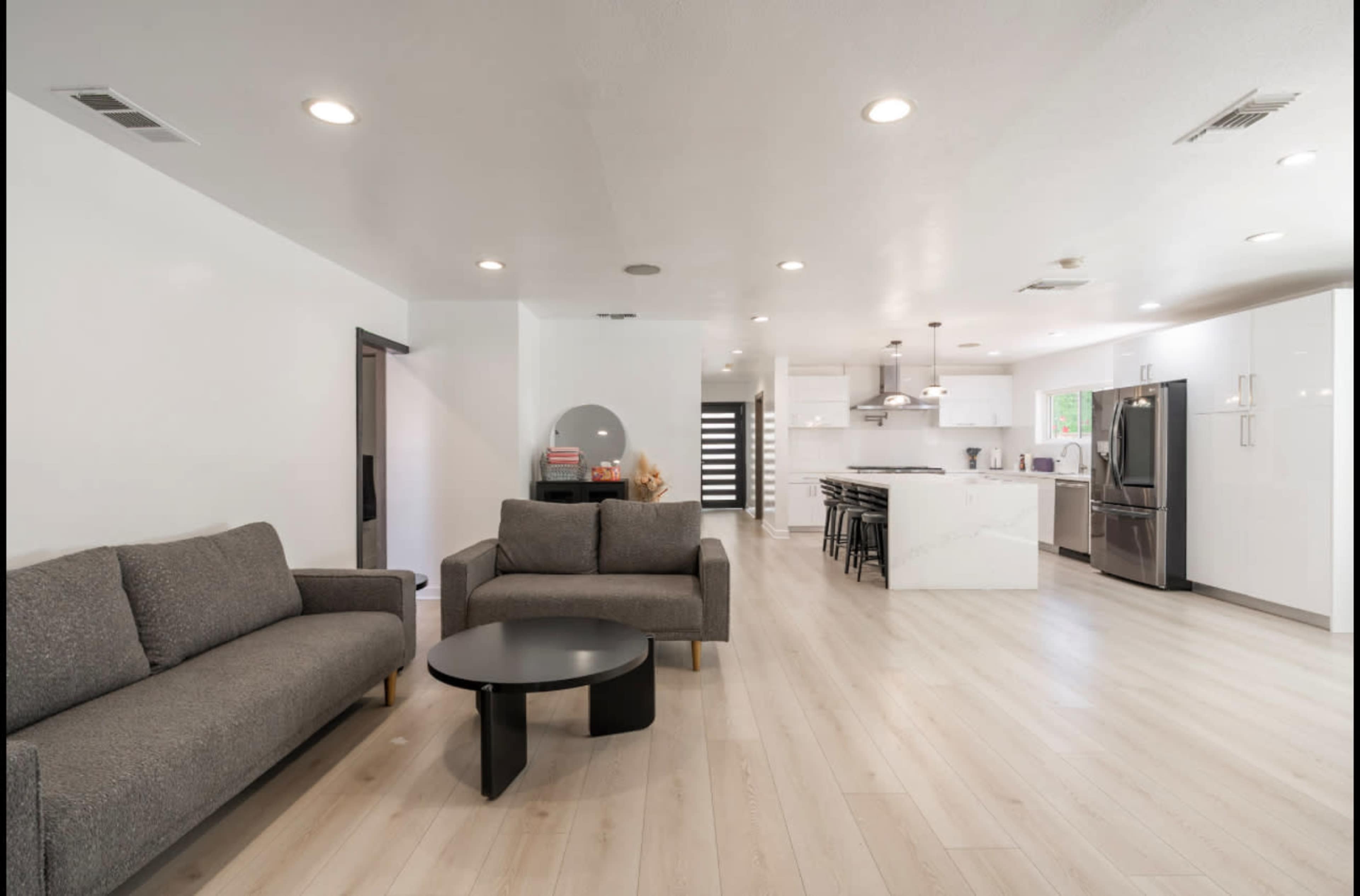 The image shows a modern, open-concept living space with a gray couch, a black coffee table, and a kitchen area featuring white cabinets and stainless steel appliances.