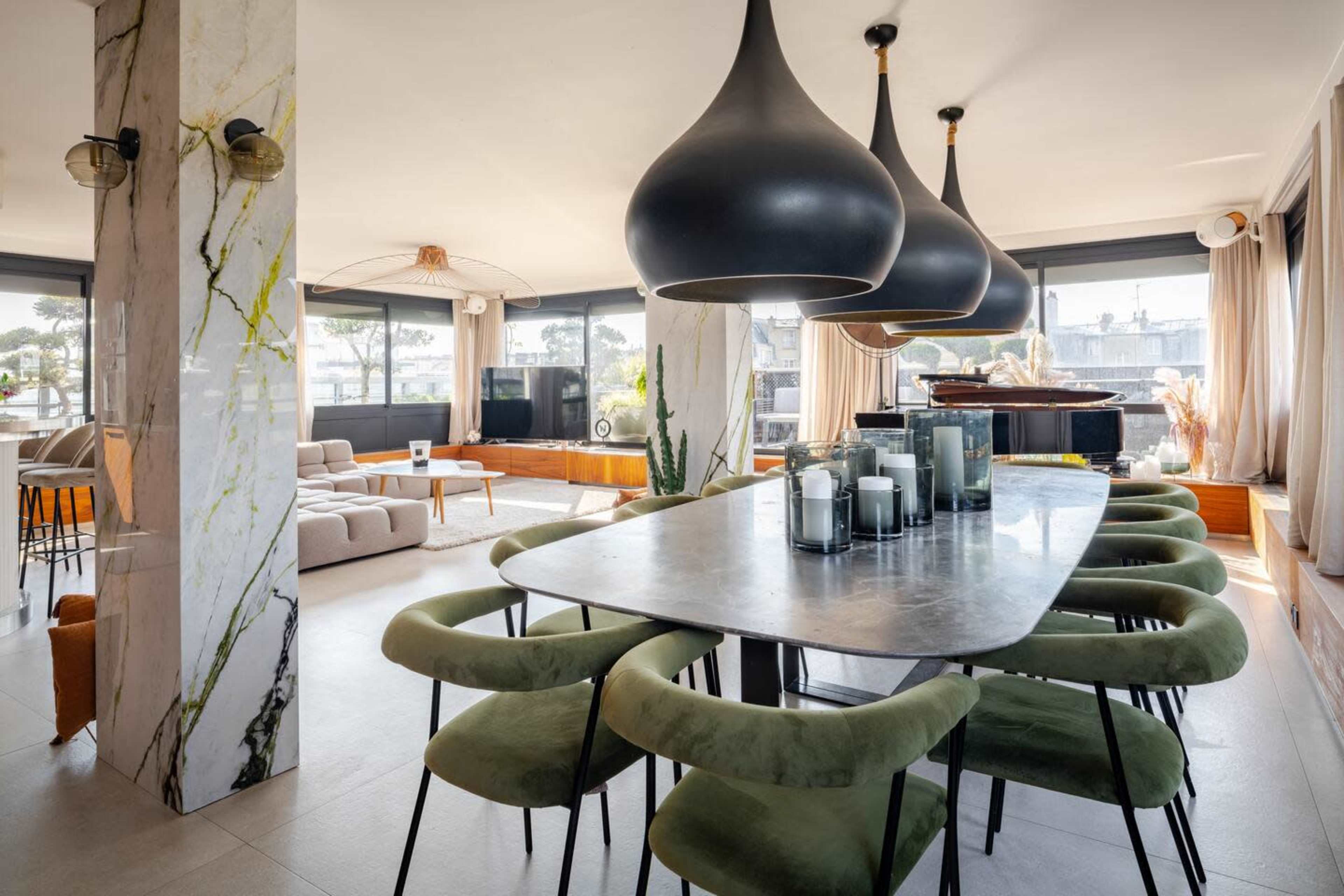 A spacious modern dining area features a large table surrounded by green chairs, with distinctive black pendant lights hanging above and large windows offering views of the outside.