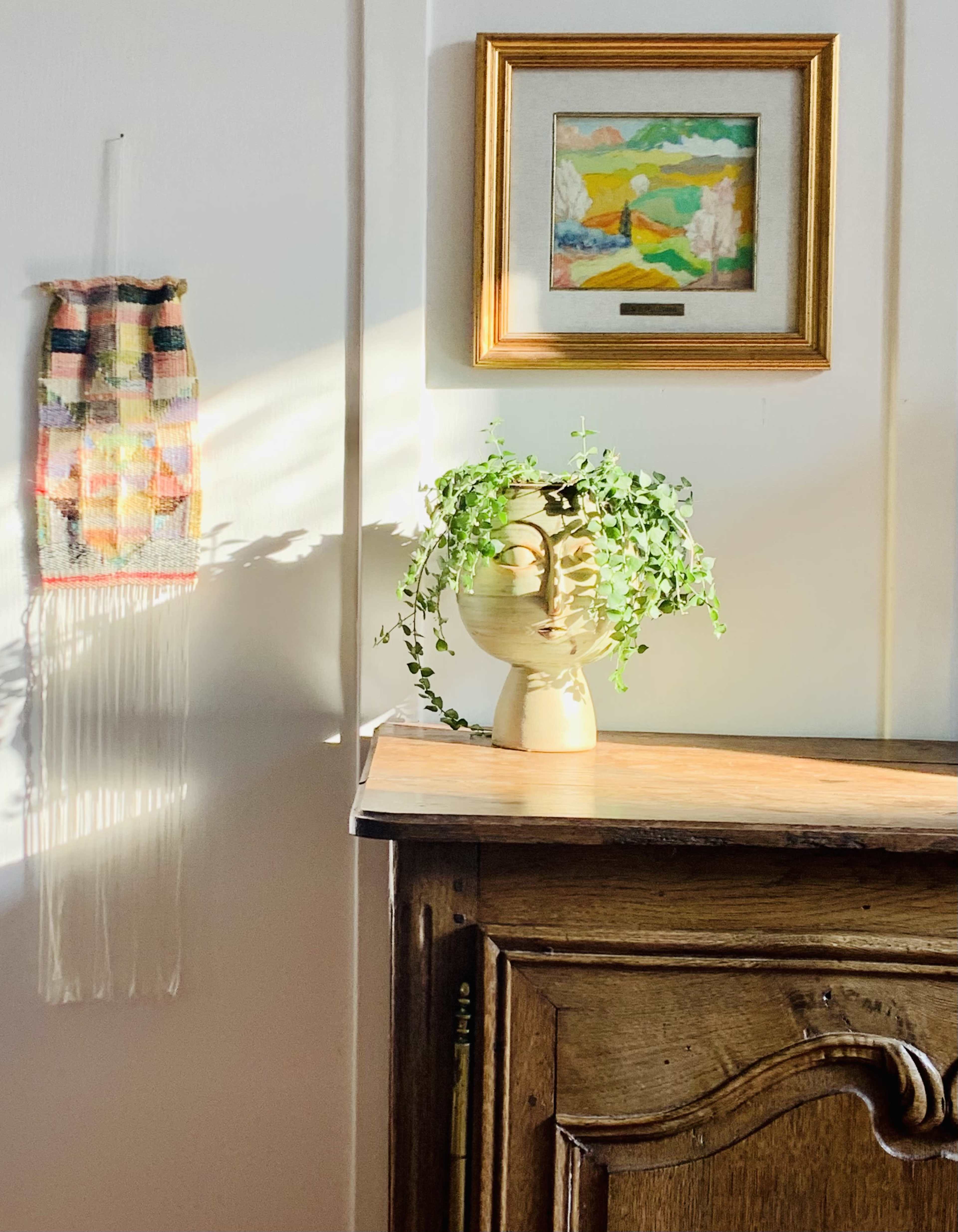 A wooden dresser is positioned beside a wall featuring a decorative textile and a framed artwork, with a plant in a vase placed on top.