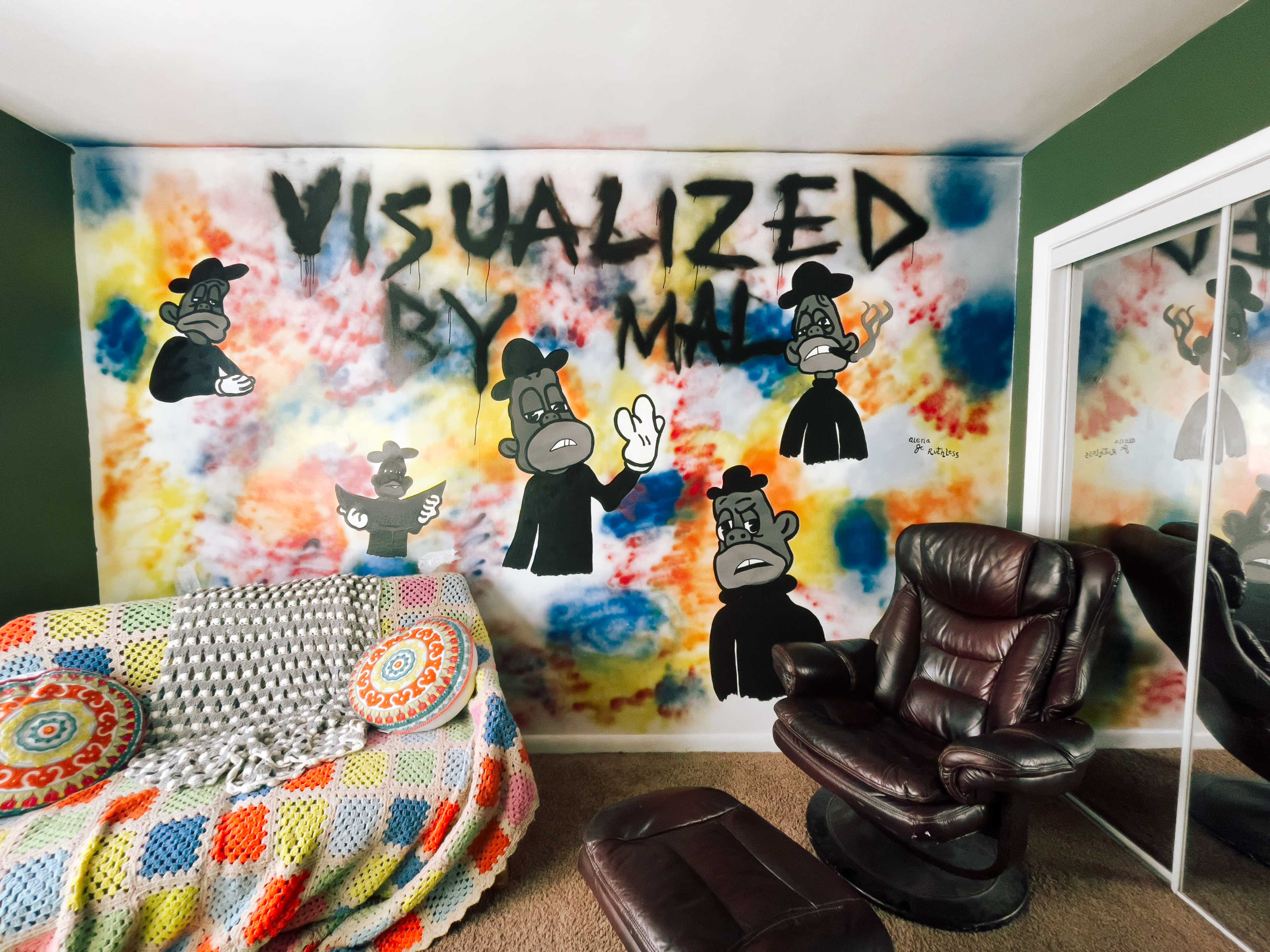 A room with a colorful graffiti mural featuring cartoon characters and the text "VISUALIZED BY MAL" on one wall, along with a patterned blanket and a brown leather chair.