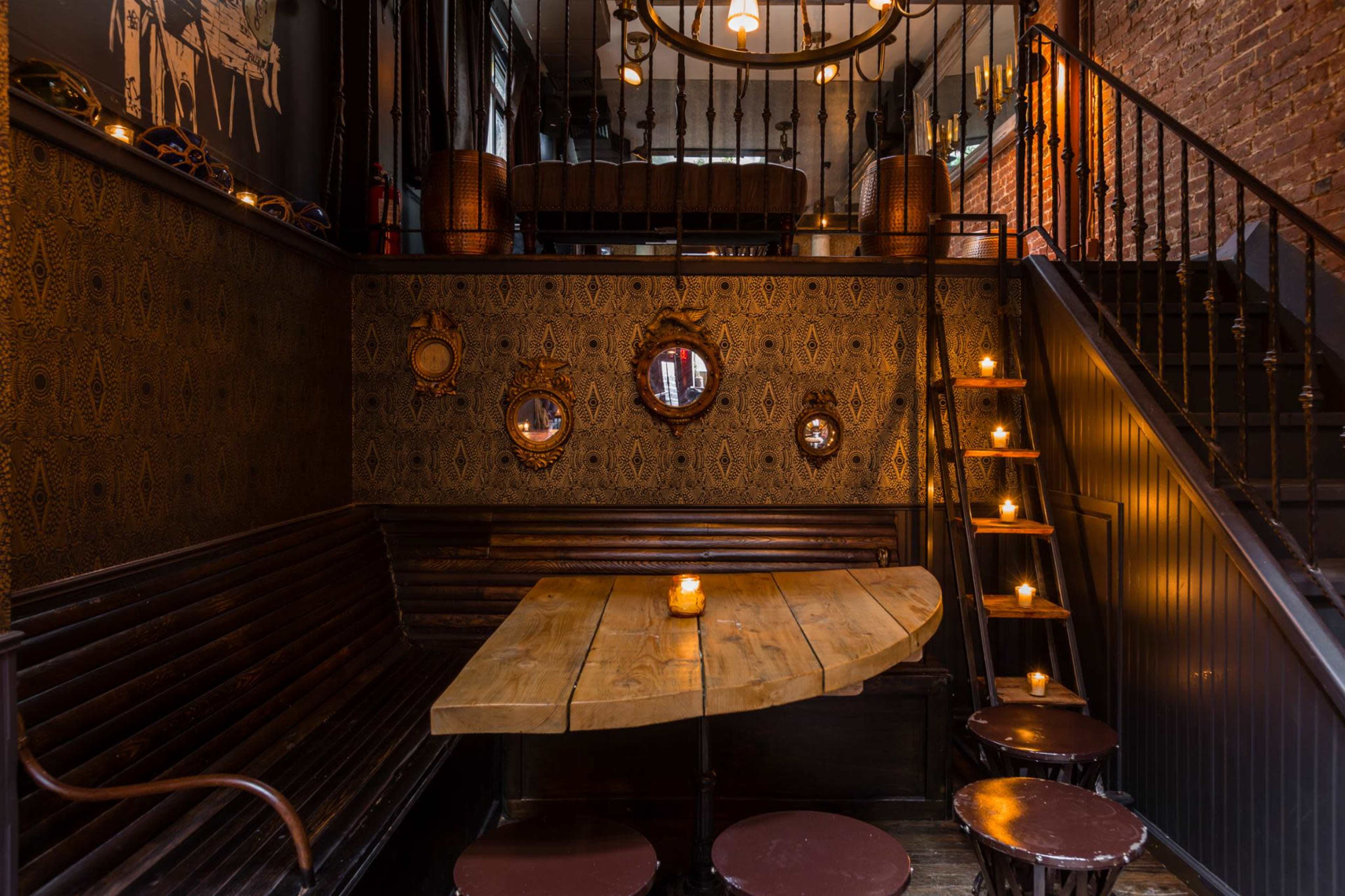 A dimly lit, eclectic interior of a bar or restaurant, featuring wooden seating, a staircase, and several small mirrors on the wall.