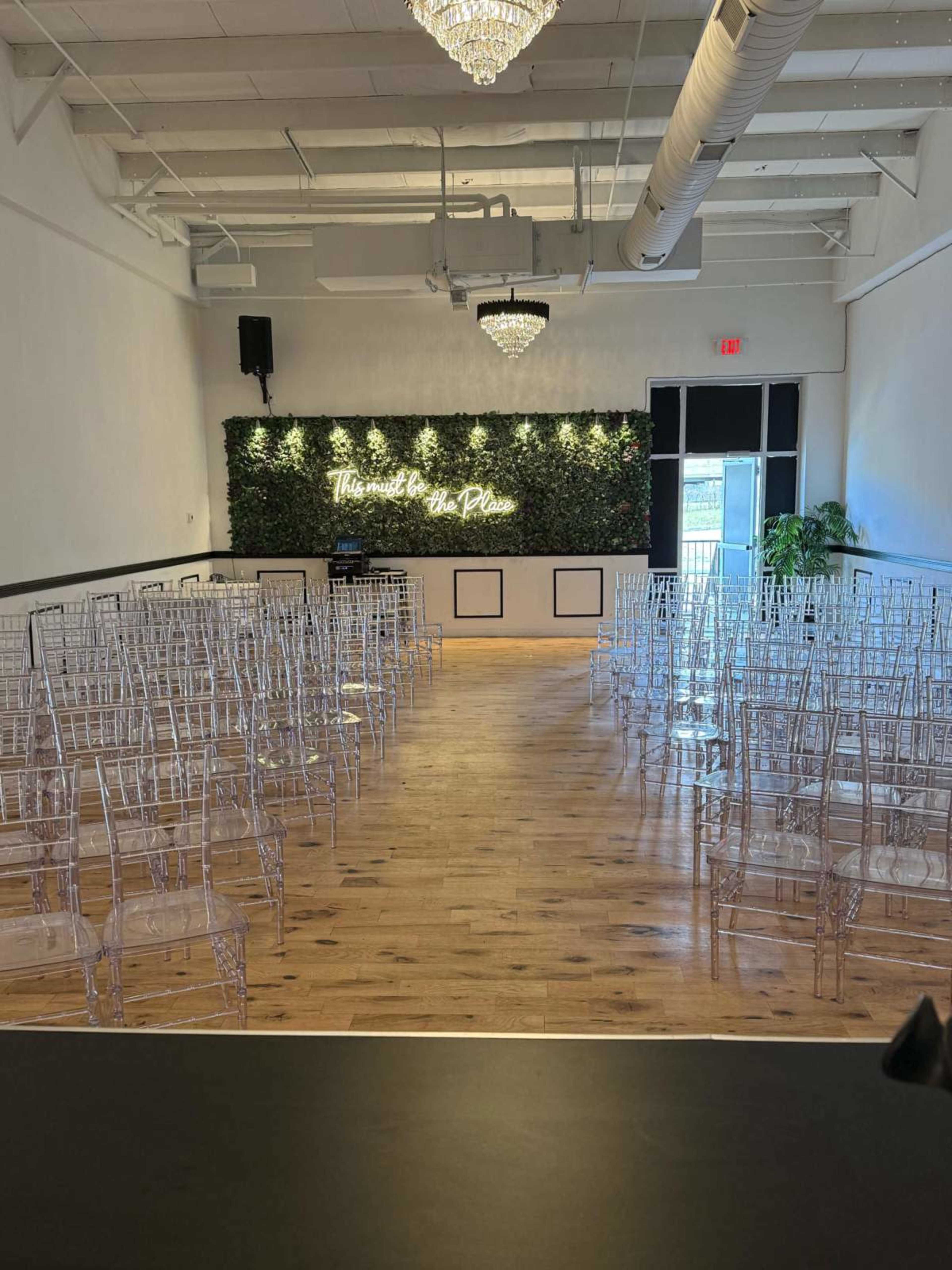 The image shows a spacious event room set up with clear acrylic chairs arranged in rows facing a greenery wall with a neon sign in the background.