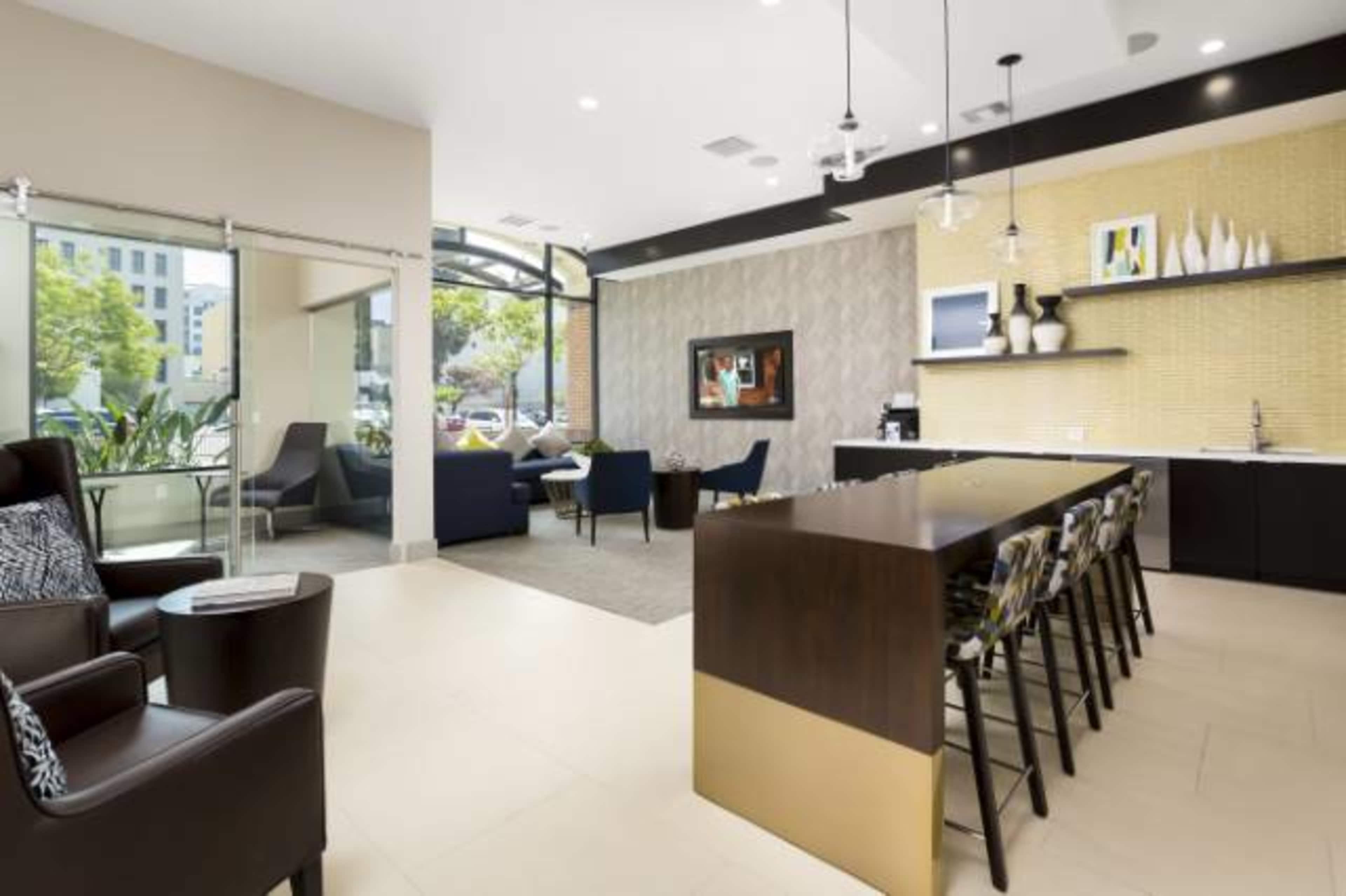 A modern lounge area featuring seating arrangements, a long bar with stools, and large windows that provide natural light.