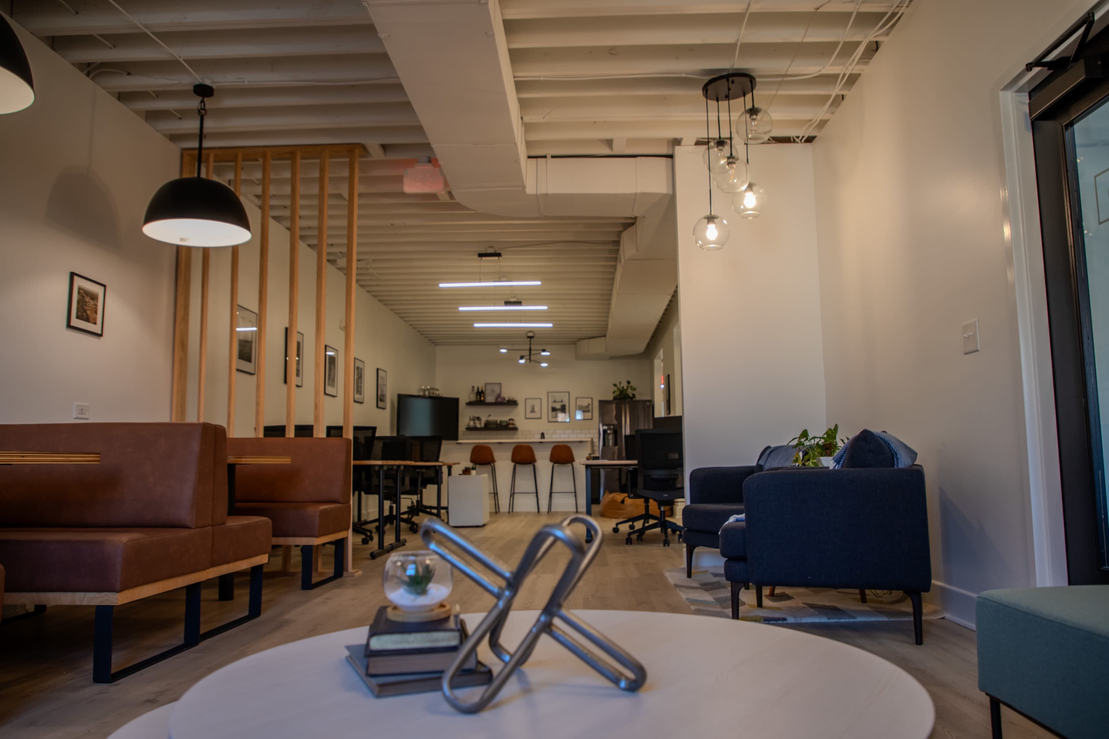 The image shows a modern office space with seating areas, a kitchen in the background, and several workstations along the wall.