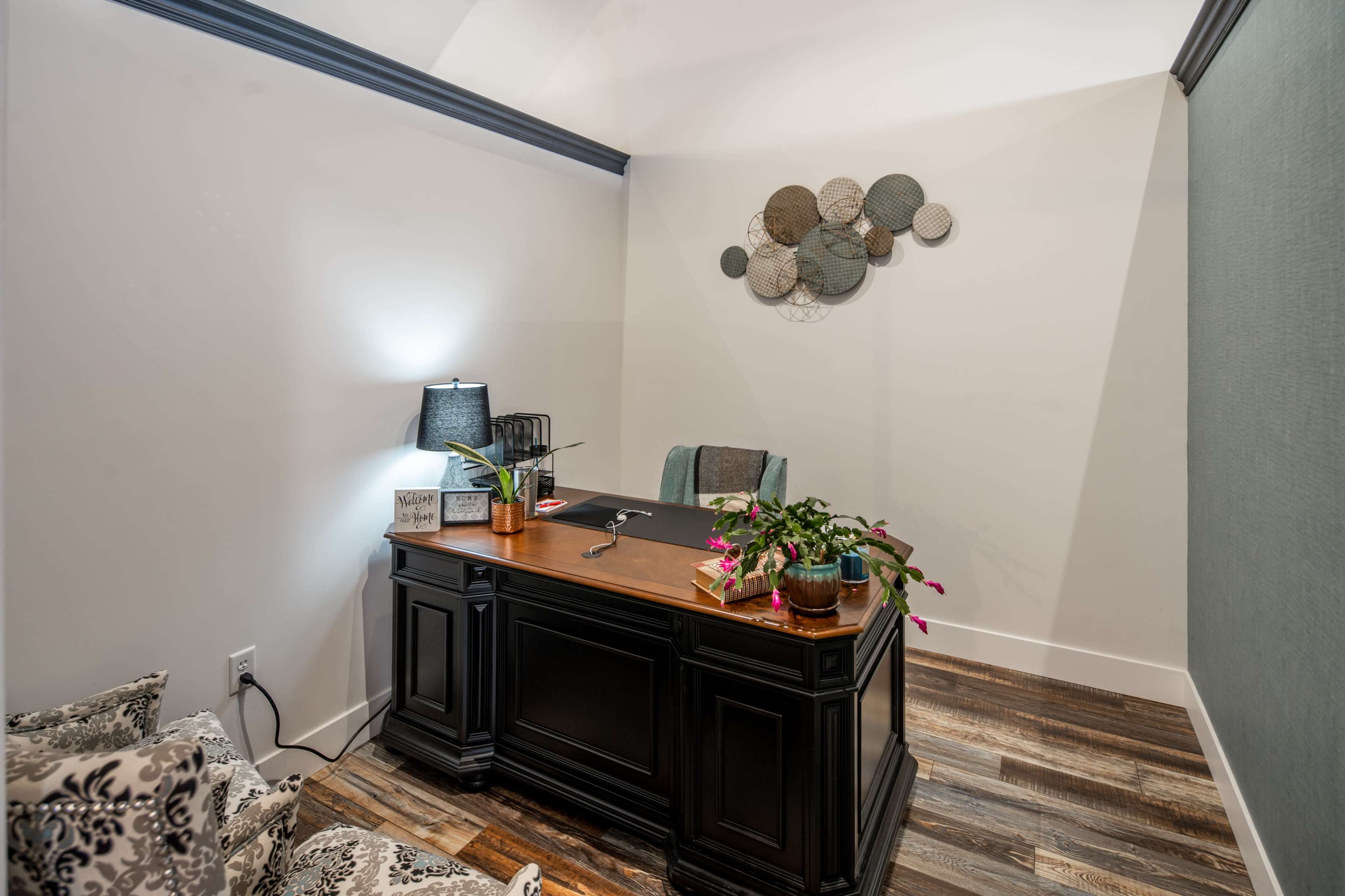 A modern office space features a dark desk, a lamp, and decorative wall art, with a plant on the desk and a cozy seating area nearby.
