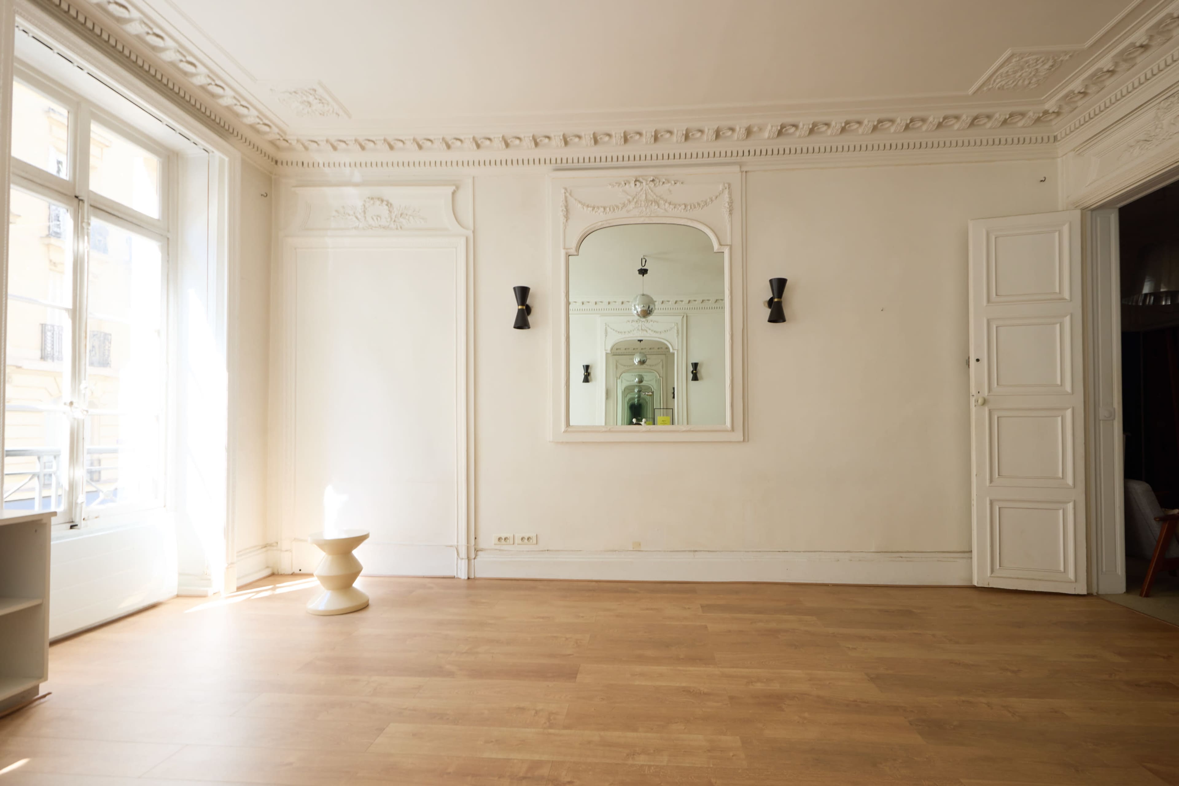 The image shows a bright, empty room with a large mirror and a small pedestal in the center, featuring tall windows that allow natural light to enter.