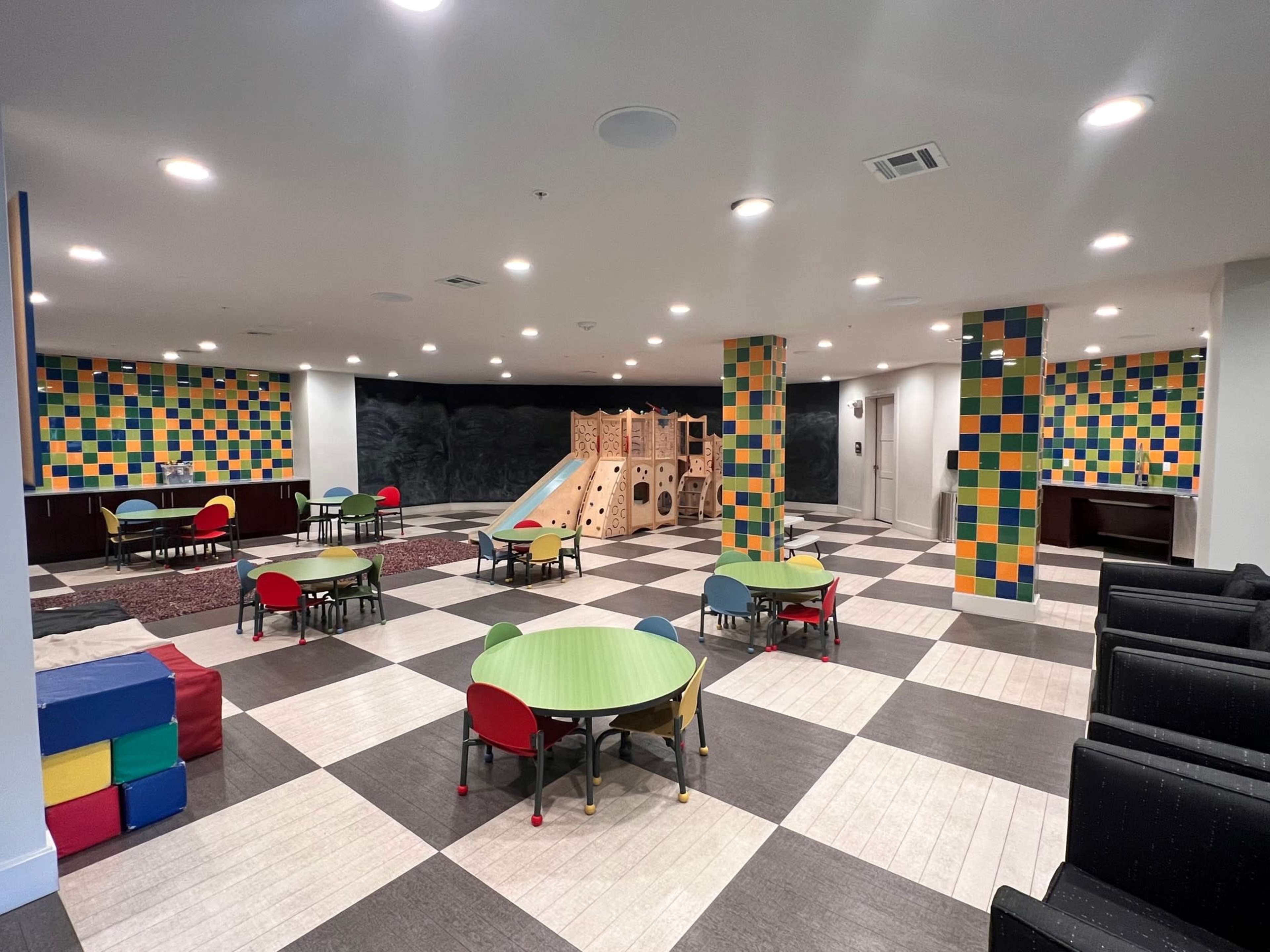 A spacious children's playroom featuring colorful walls, a small playground structure, several round tables with chairs, and a checkered floor pattern.