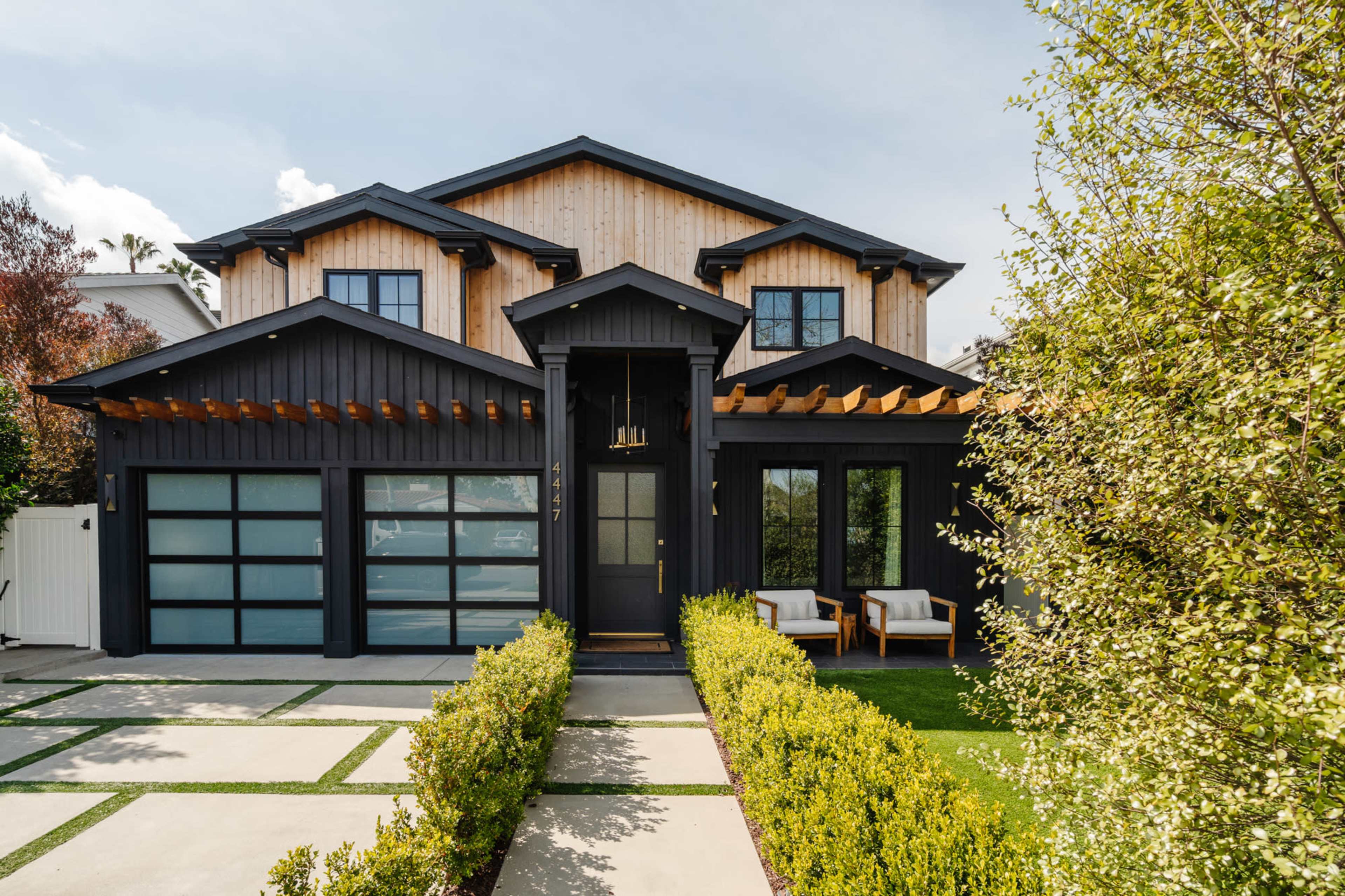 A modern black and wood house with large windows, a front porch, and a neatly landscaped pathway lined with green hedges.