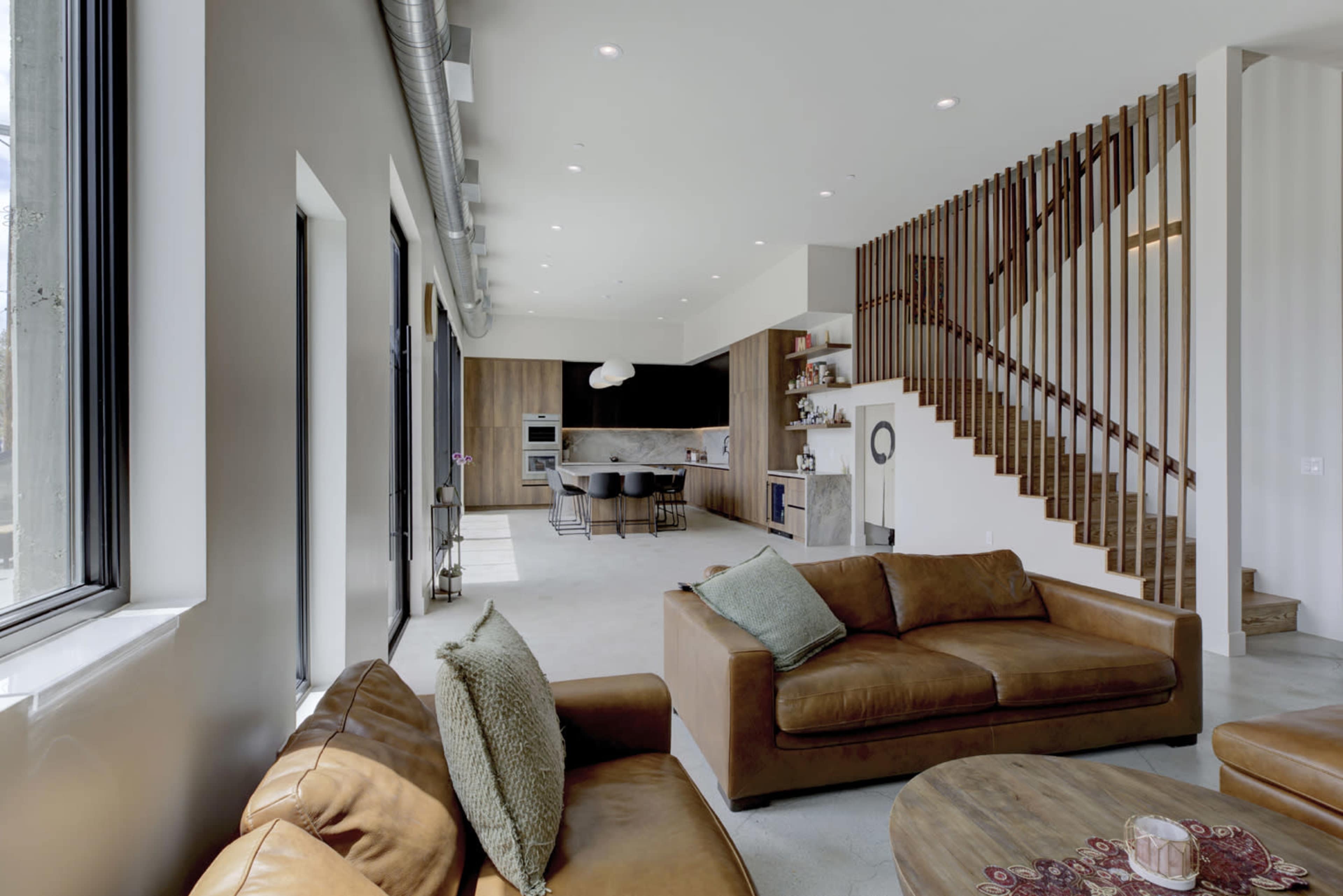 The image shows a modern, open-concept living space with a kitchen and staircase, featuring leather sofas and large windows that allow natural light to enter.