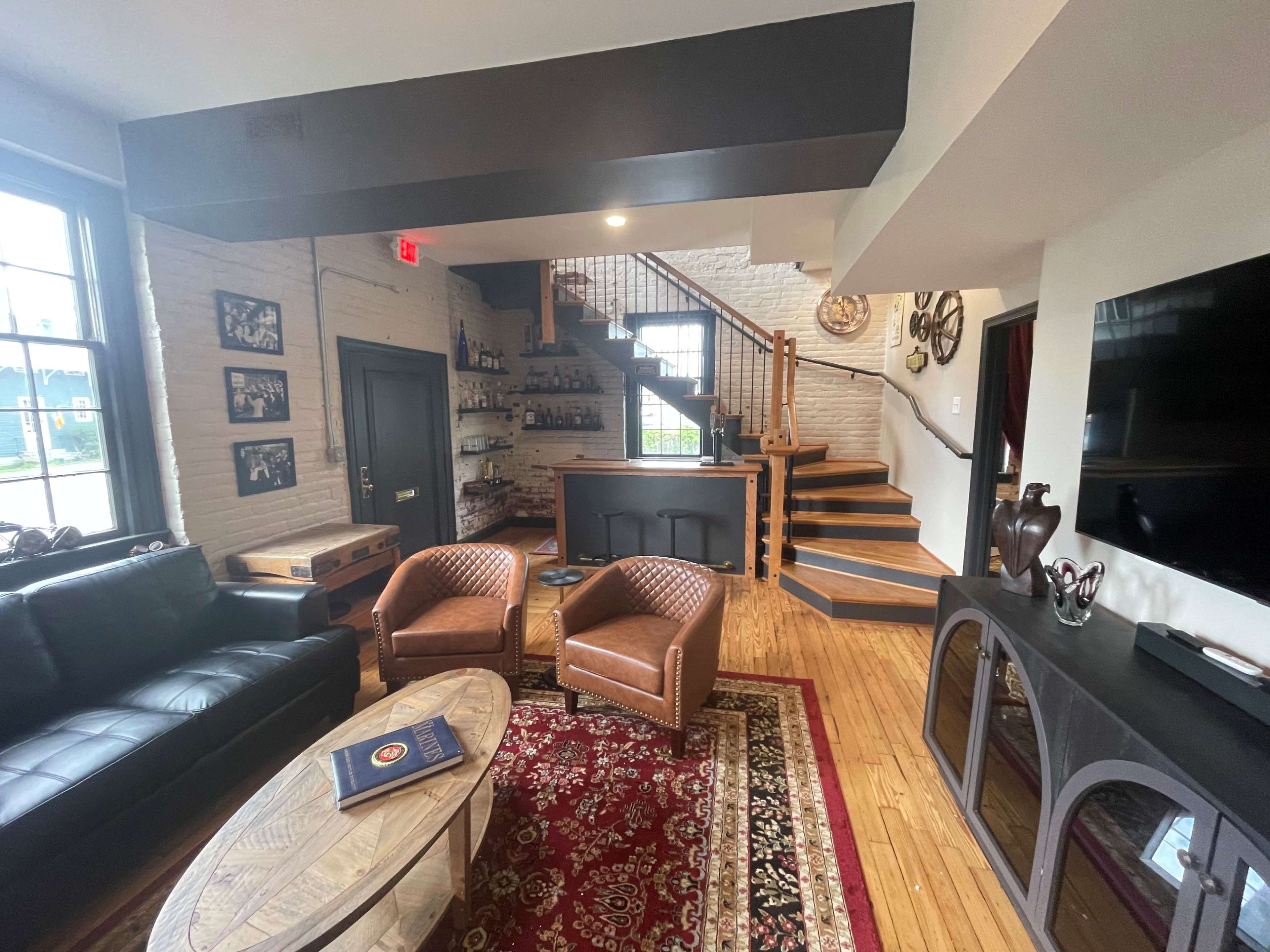 The image shows a cozy interior space featuring a black leather sofa, two brown armchairs, a round coffee table, a staircase, and a bar area in a well-lit room with wooden flooring.
