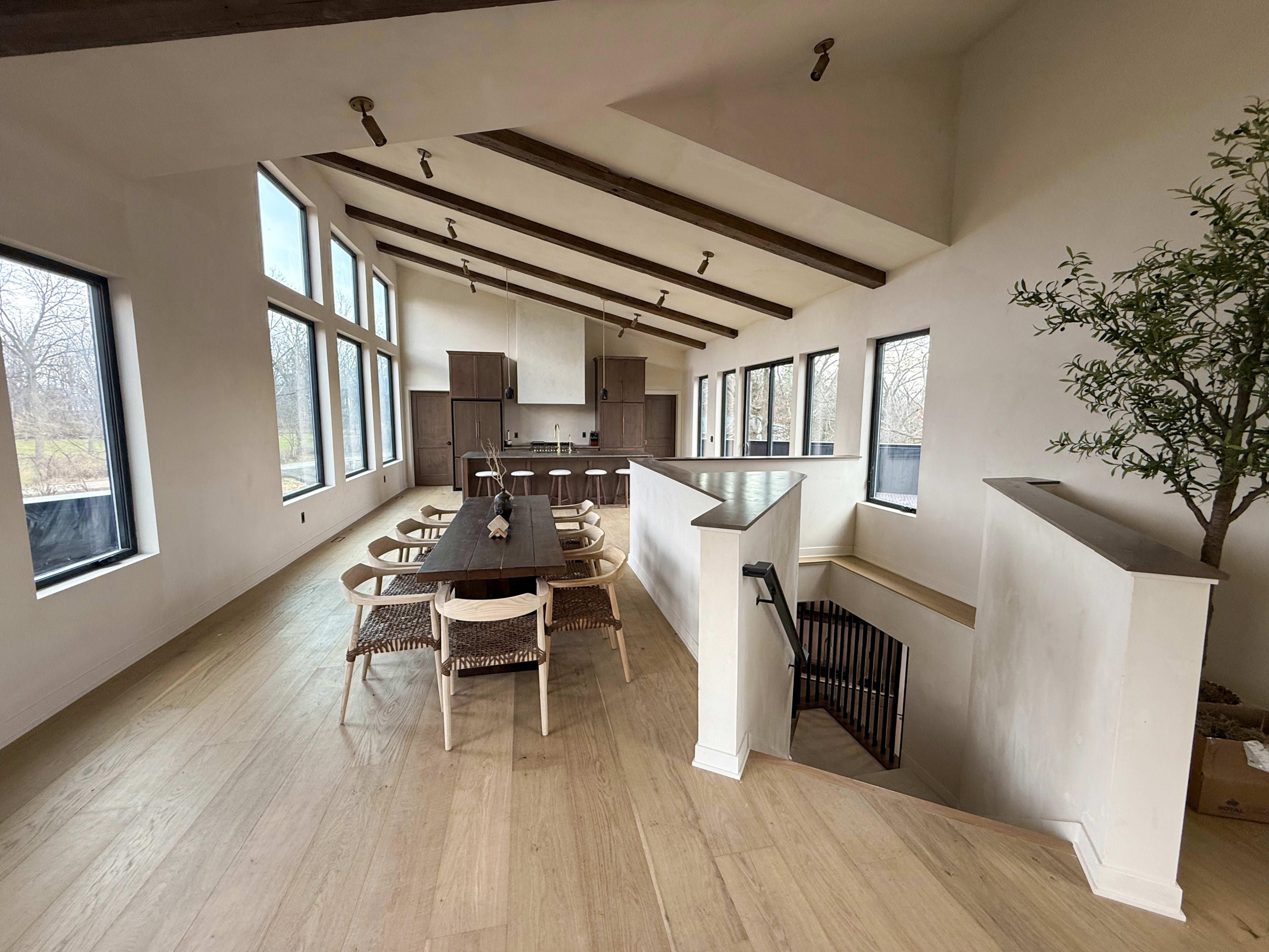 The image shows a spacious, modern open-plan dining area with a long table, surrounded by chairs, and a kitchen in the background featuring large windows and exposed wooden beams.