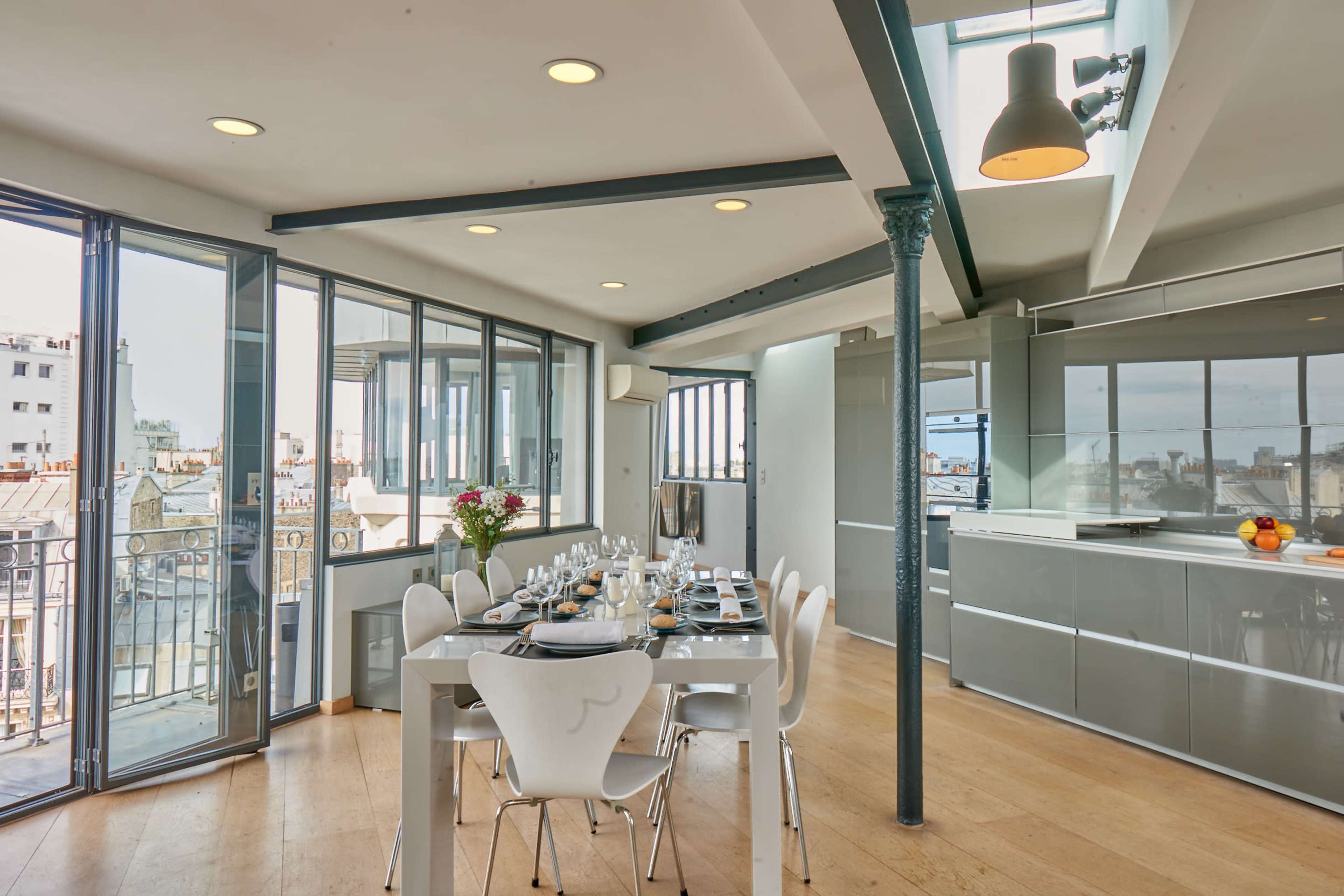A modern dining area features a large table set for a meal, surrounded by windows that provide views of the city.