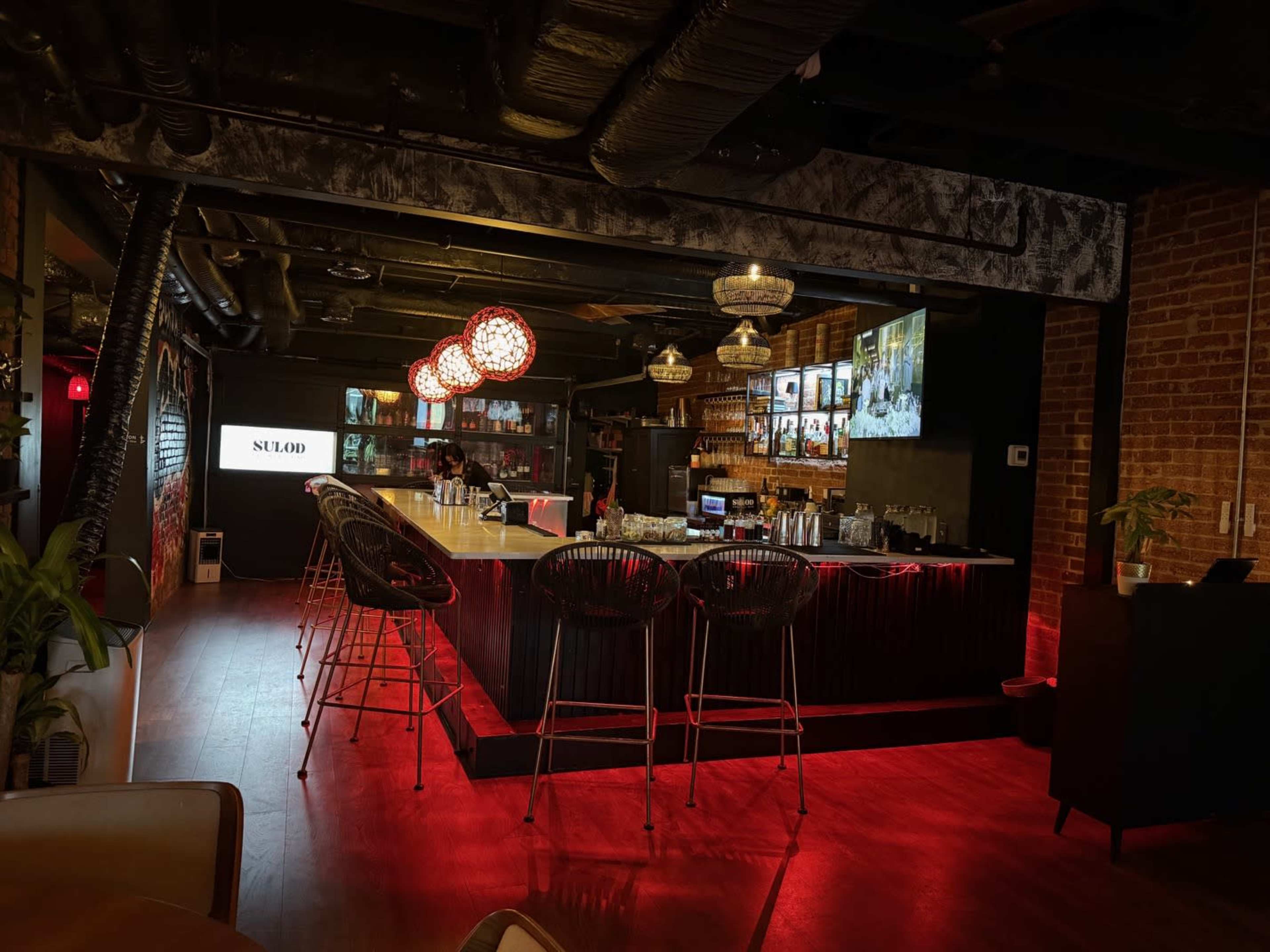 The image shows a modern bar interior featuring high stools, a long counter with various bottles, and decorative lighting in a dimly lit space.