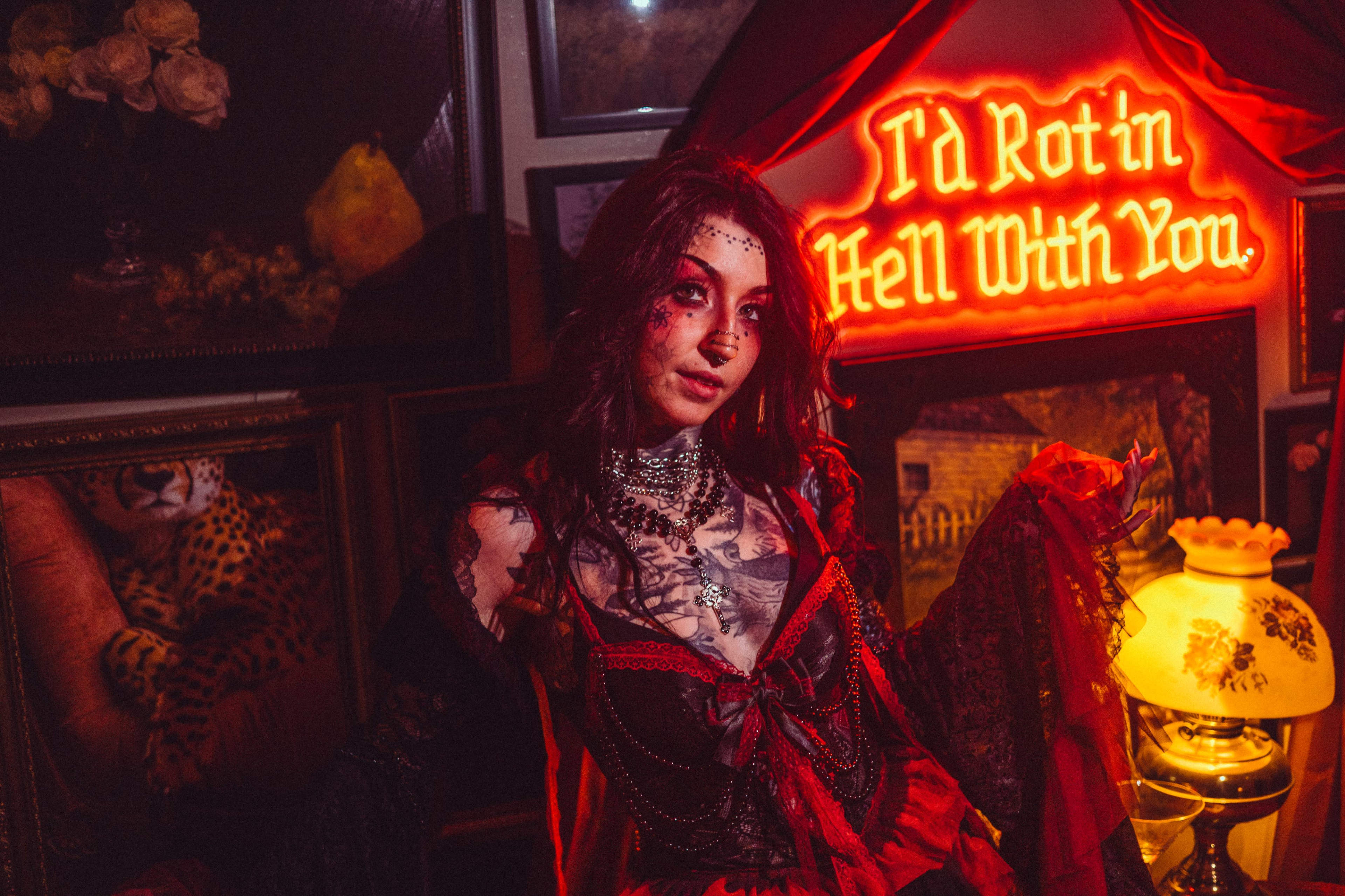 A woman in gothic attire poses in front of a neon sign that reads, "I’d Rot in Hell With You," surrounded by various artworks and decorations.