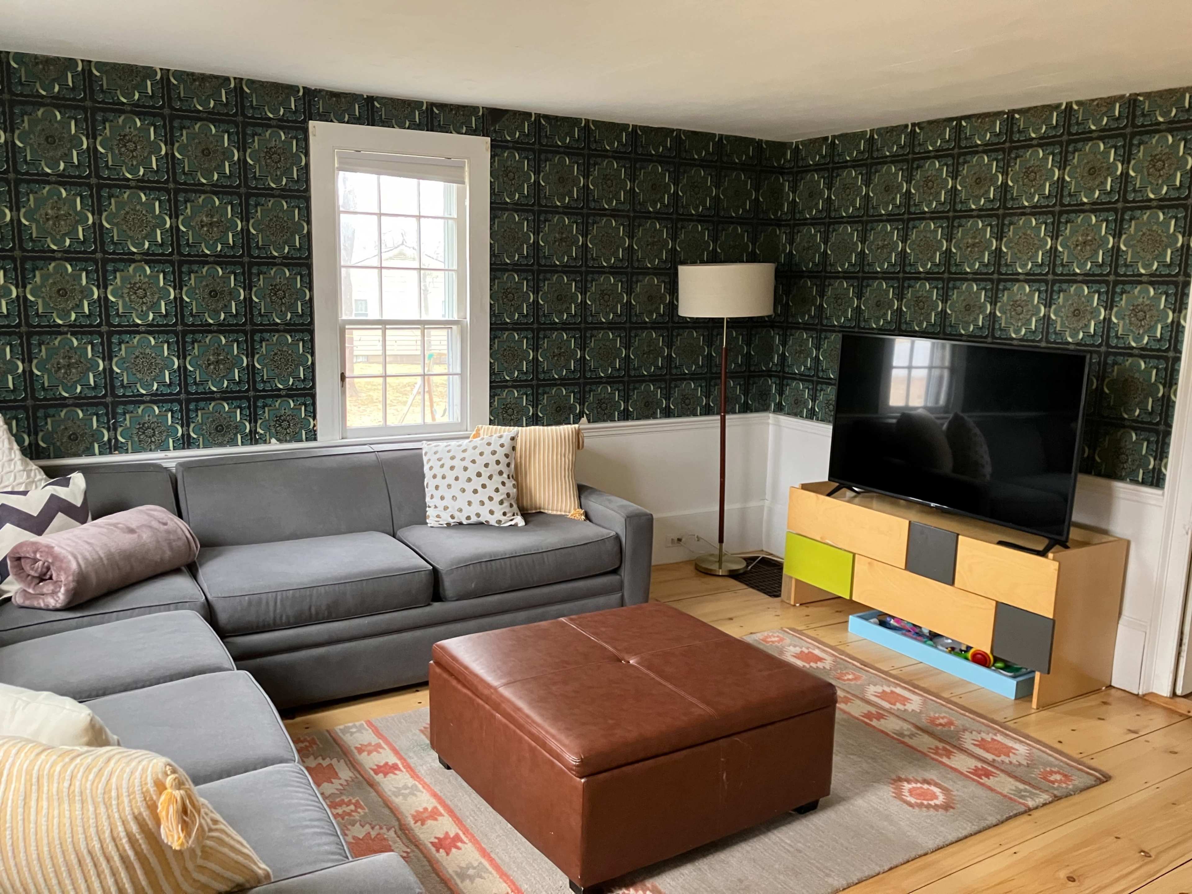 A living room features a gray sectional sofa, a brown ottoman, and a television on a low cabinet against a patterned wallpapered wall.