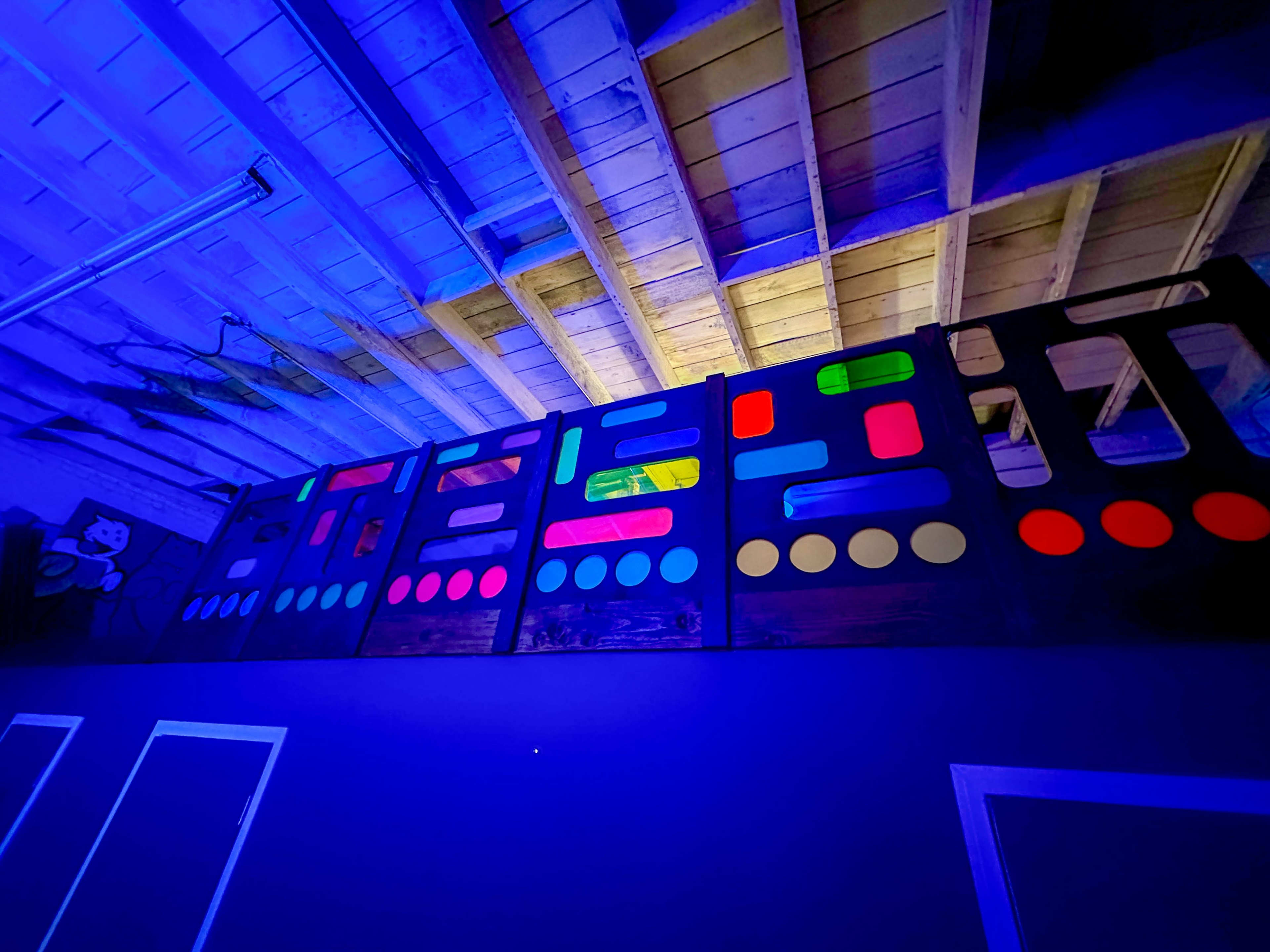 A colorful abstract mural made of various shapes and bright colors mounted on a wooden structure under a blue-lit ceiling.