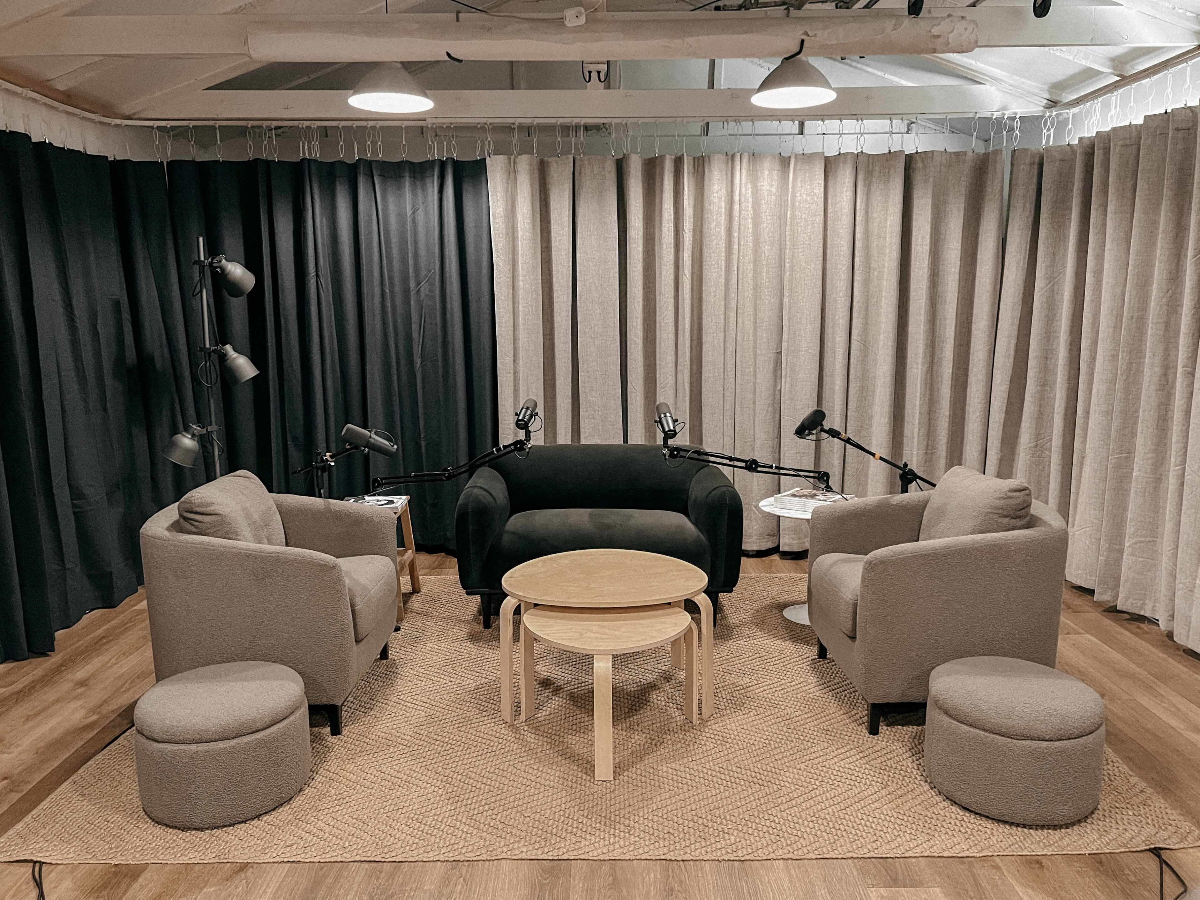 A cozy podcast studio features a central table surrounded by four armchairs and soft seating, with lamps and microphones positioned nearby.