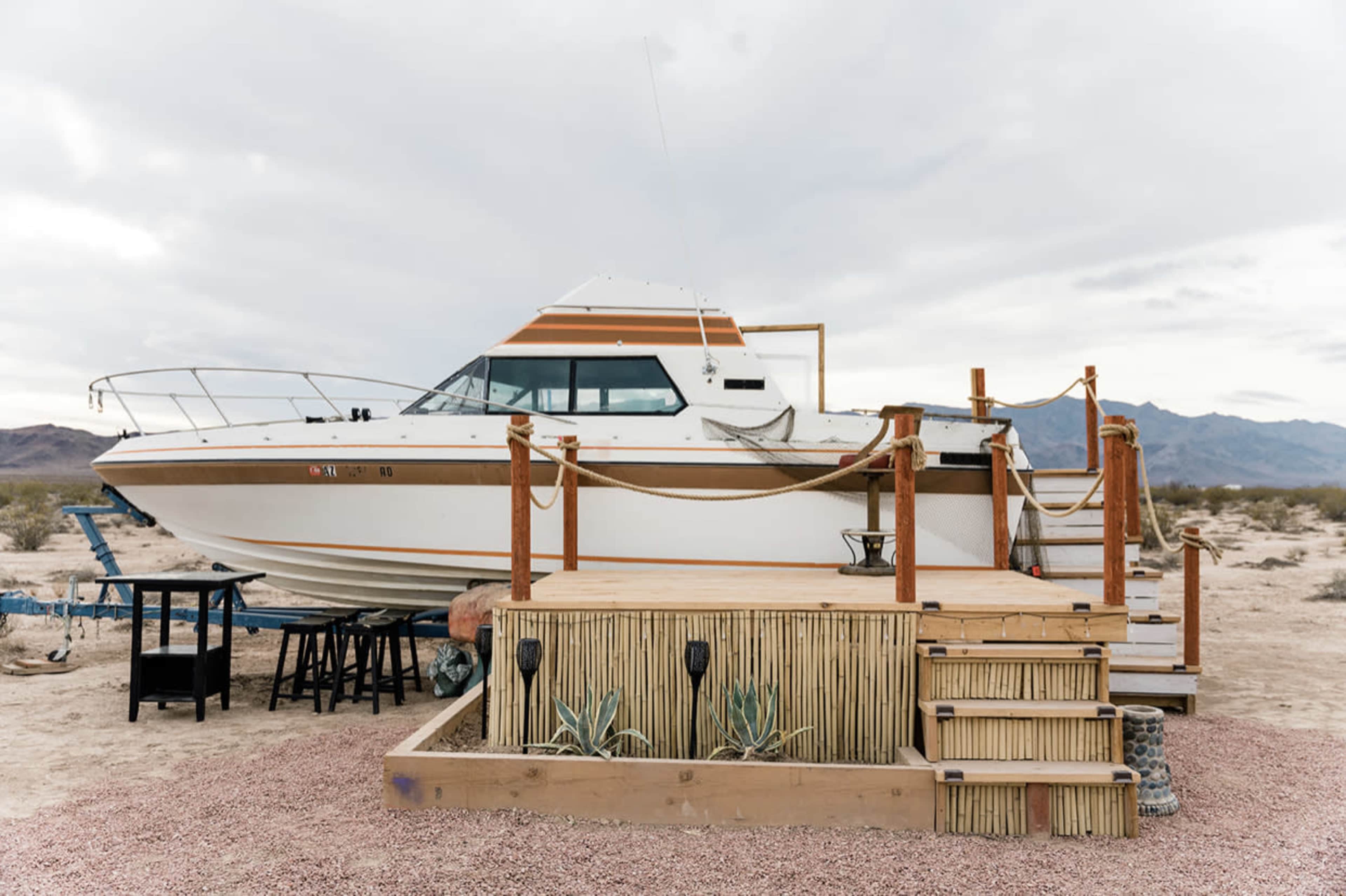 A boat is perched on a raised wooden platform in a desert landscape, with a few tables nearby and mountains visible in the background.