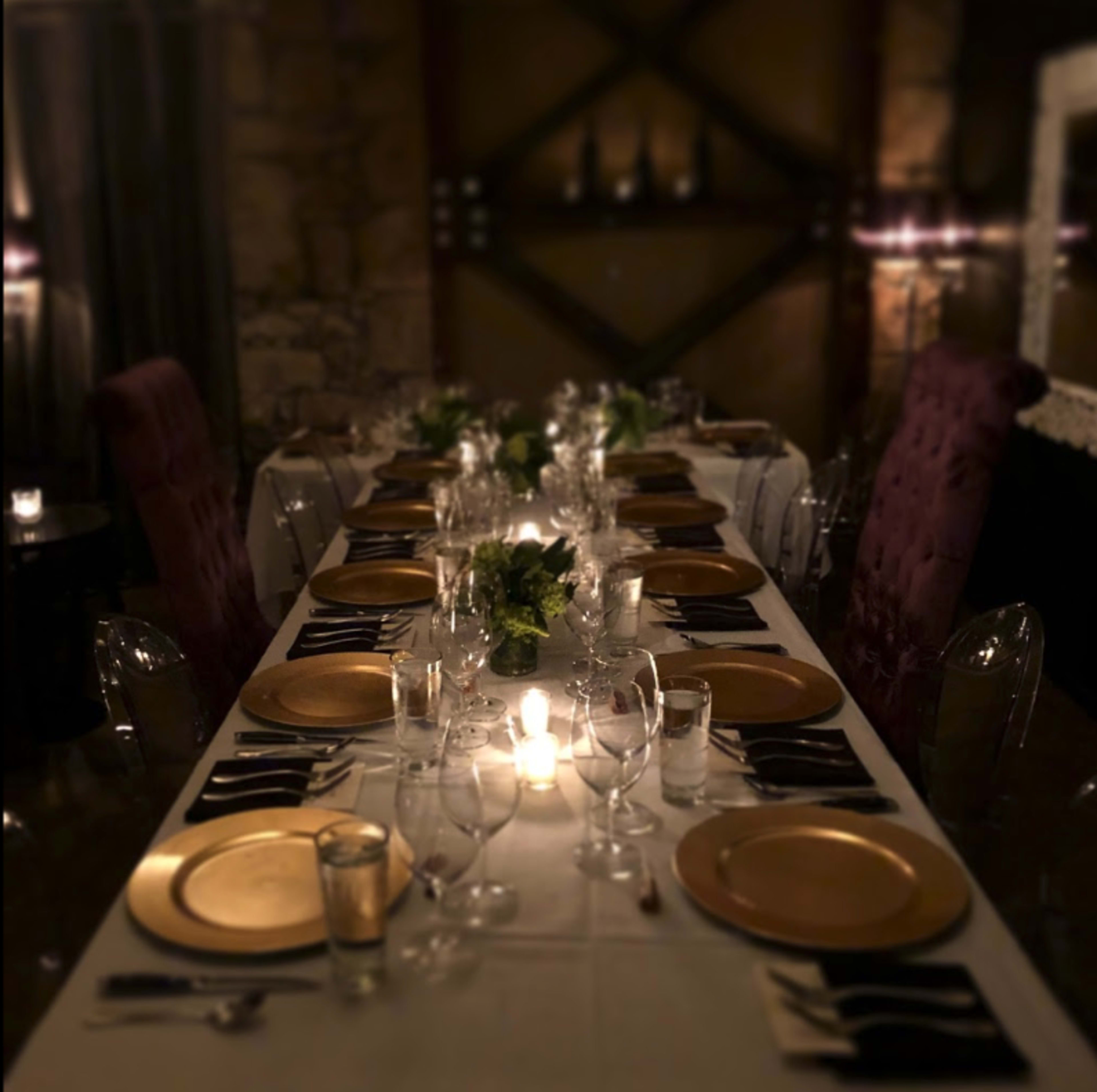 A long dining table is set with gold plates, glassware, and small green centerpieces, illuminated by candlelight in a dimly lit room.