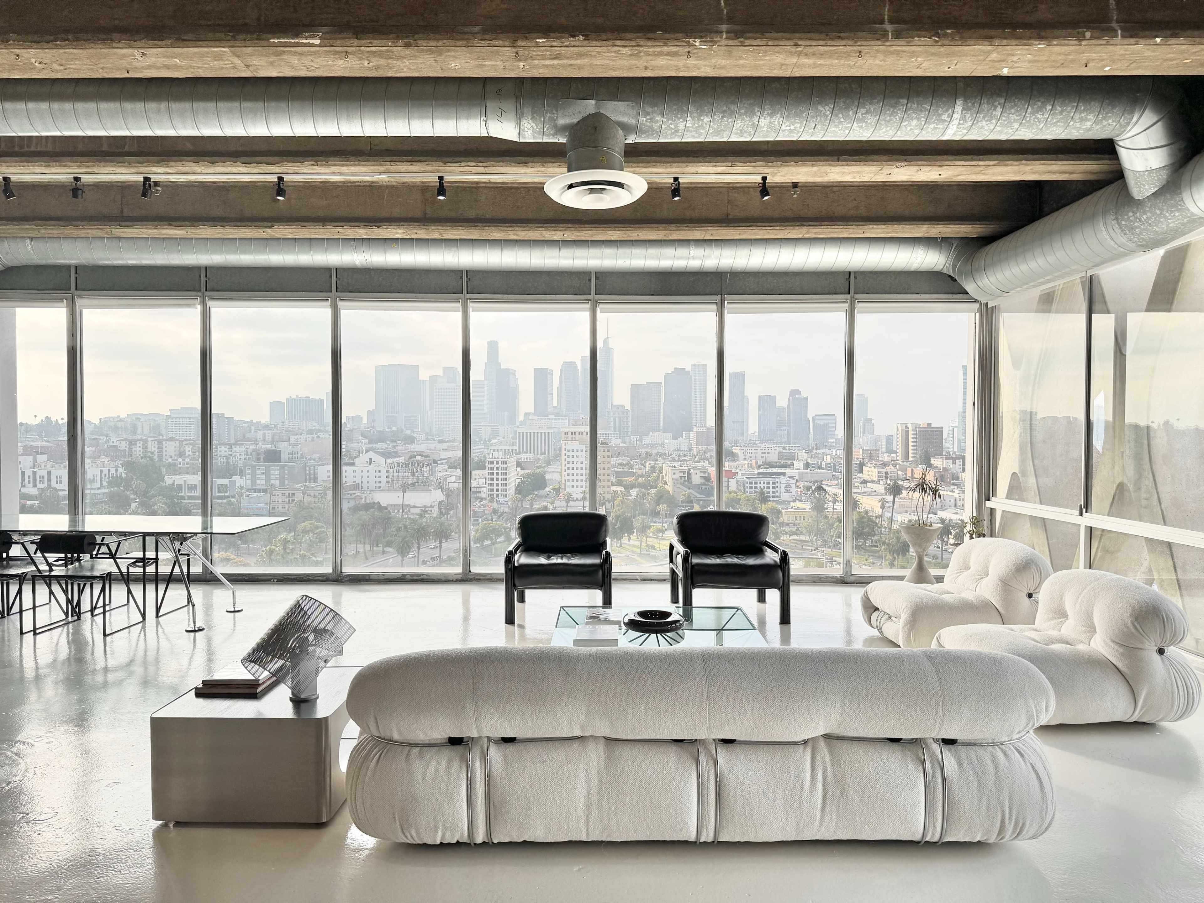 A modern living space features a white sofa and black chairs, with large windows showcasing a city skyline.