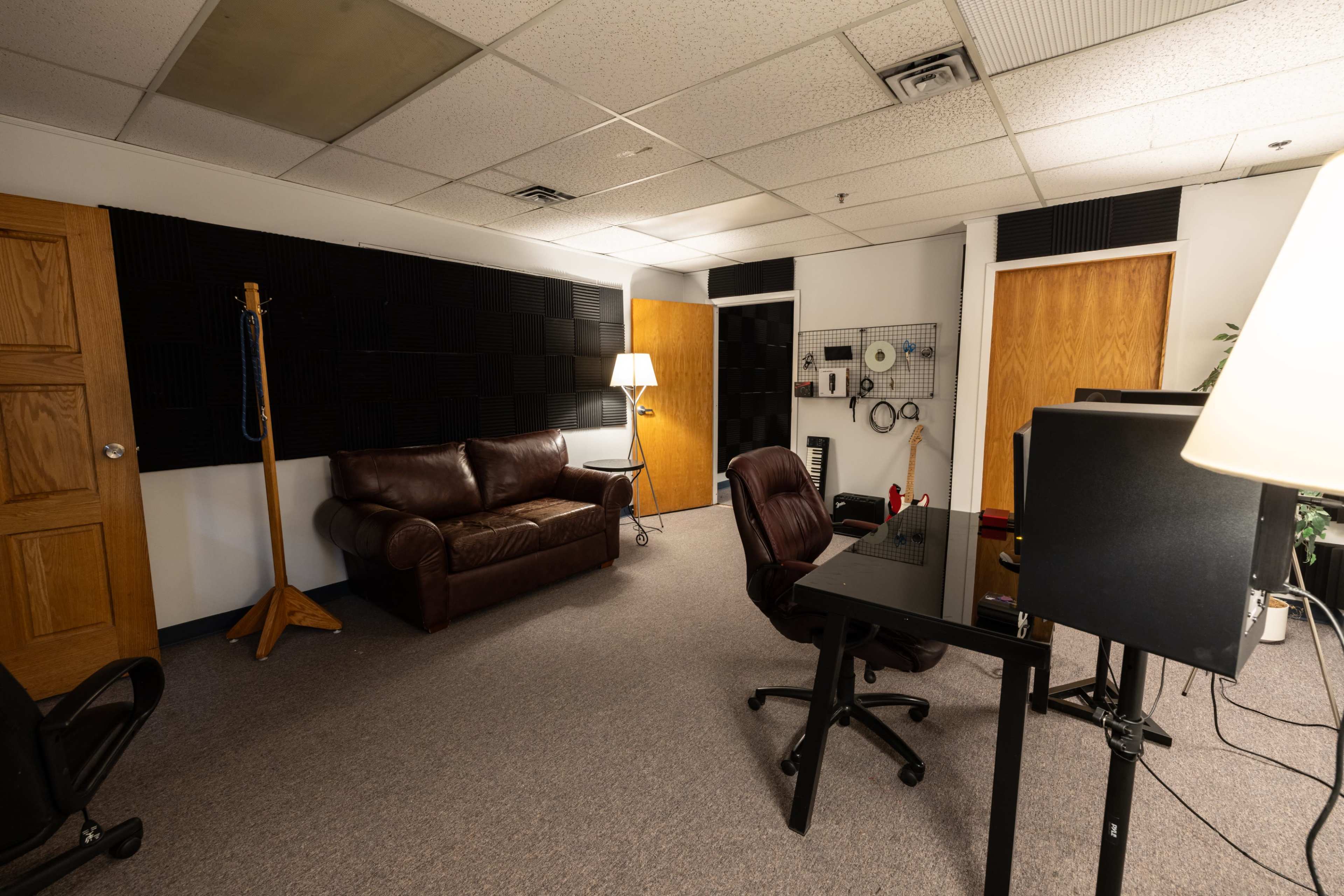 The image shows a sparsely furnished room with a brown leather couch, a desk and chair, and soundproofing panels on the walls.