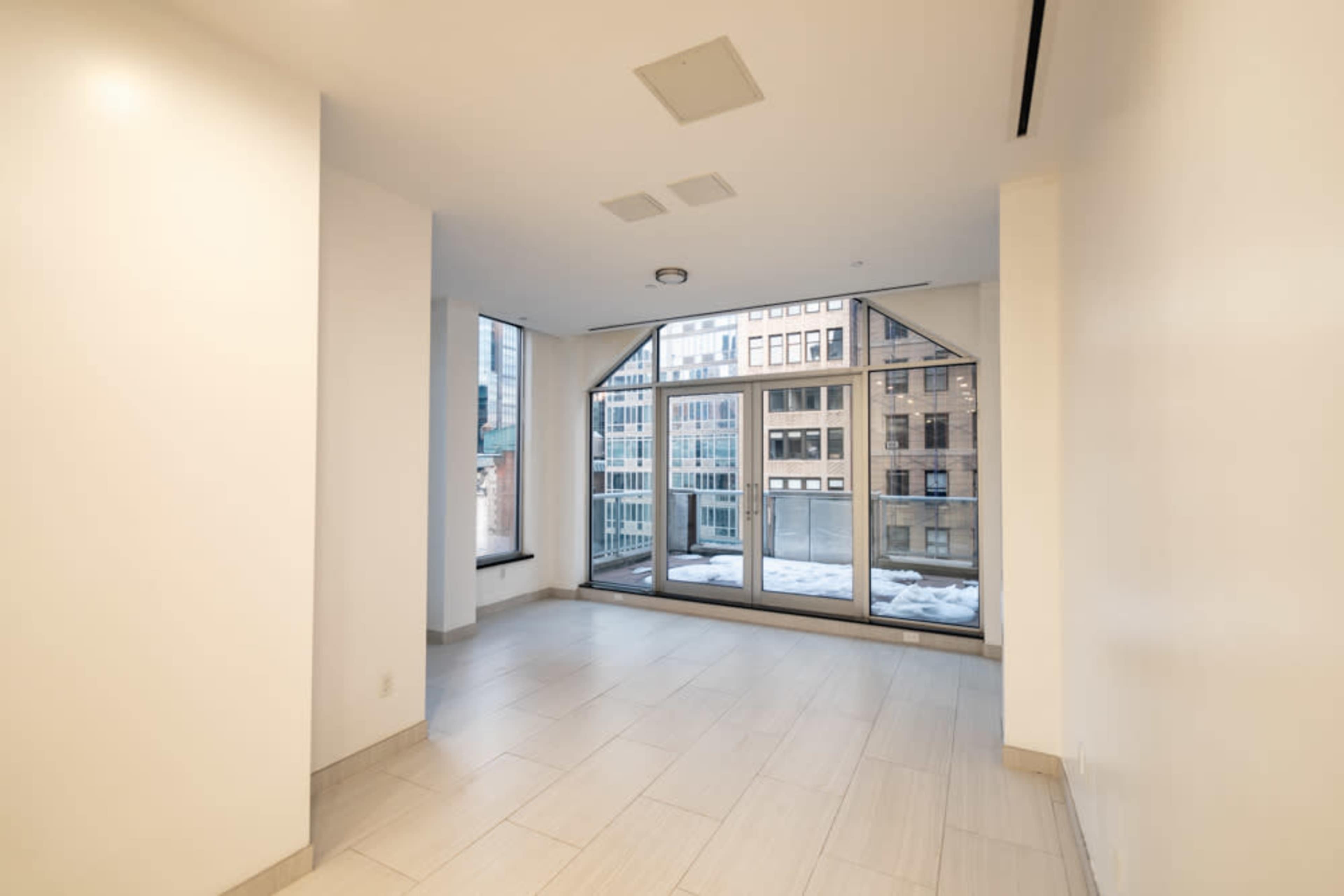 The image shows an empty room with large windows that overlook a cityscape, featuring a tiled floor and smooth white walls.