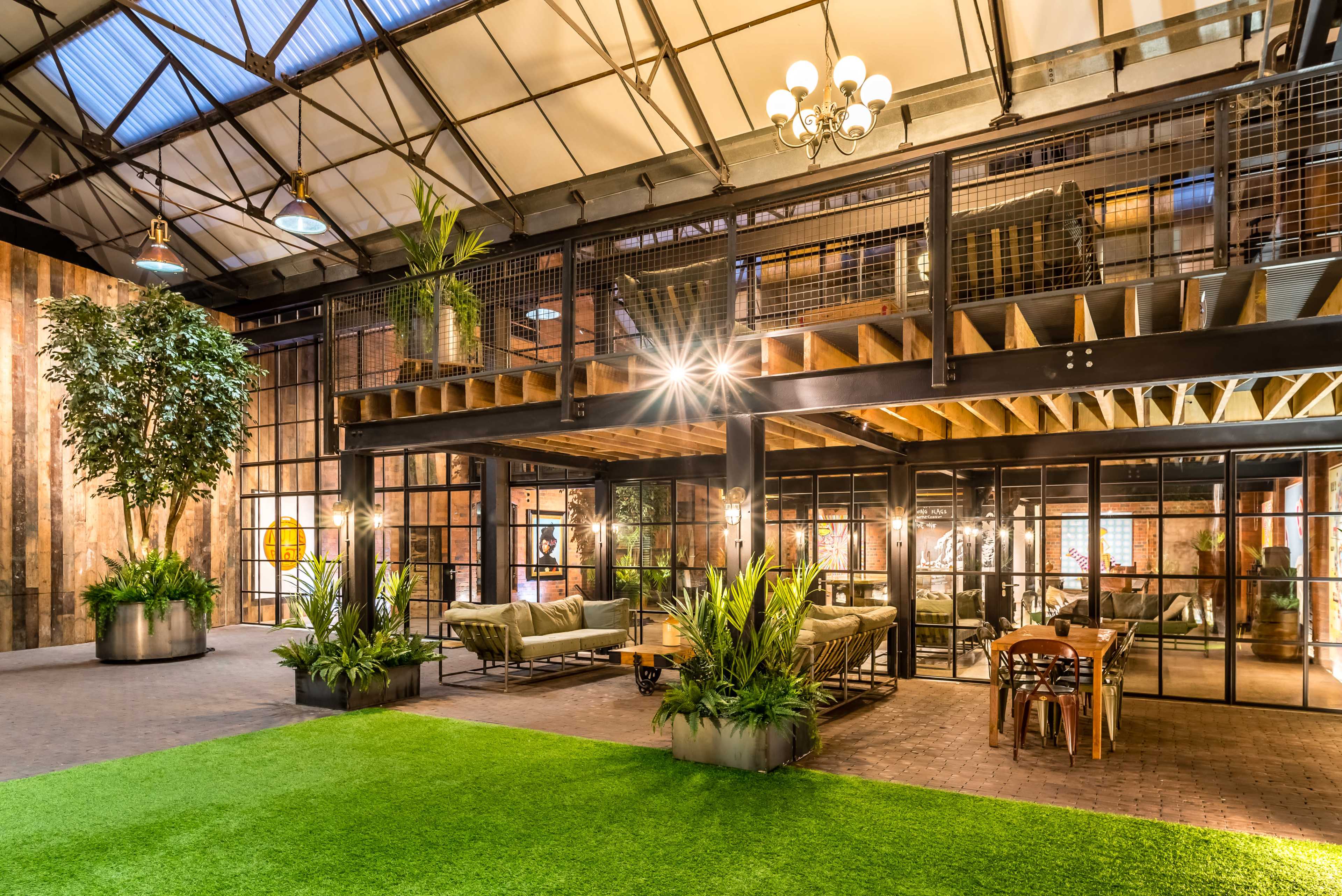 An industrial-style interior space featuring a large open area with a green lawn, plants, and metal and wooden furnishings, illuminated by multiple light fixtures.