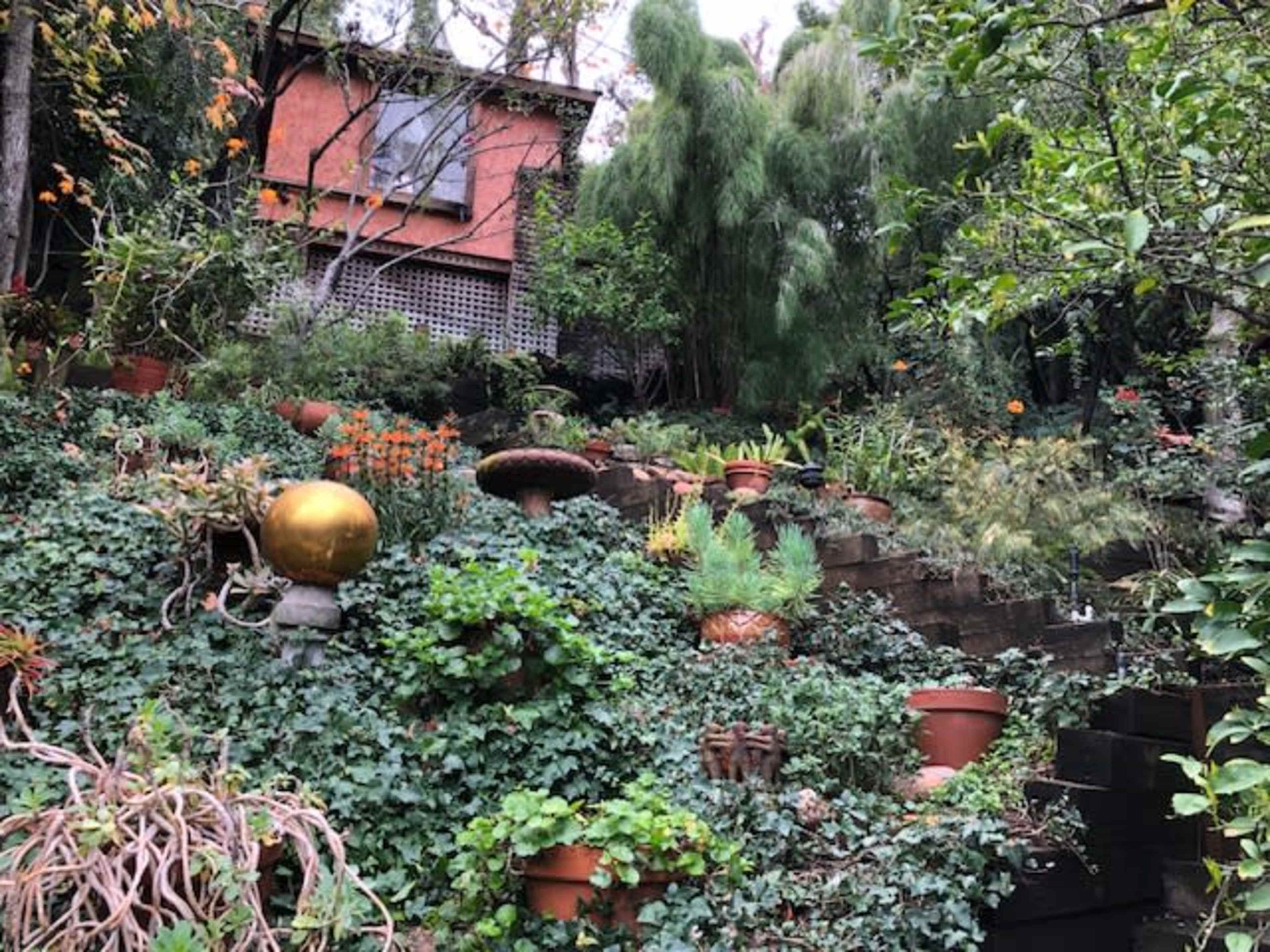 A garden hill with terraced steps features various potted plants and a golden orb sculpture, all set against a backdrop of greenery and a house above.
