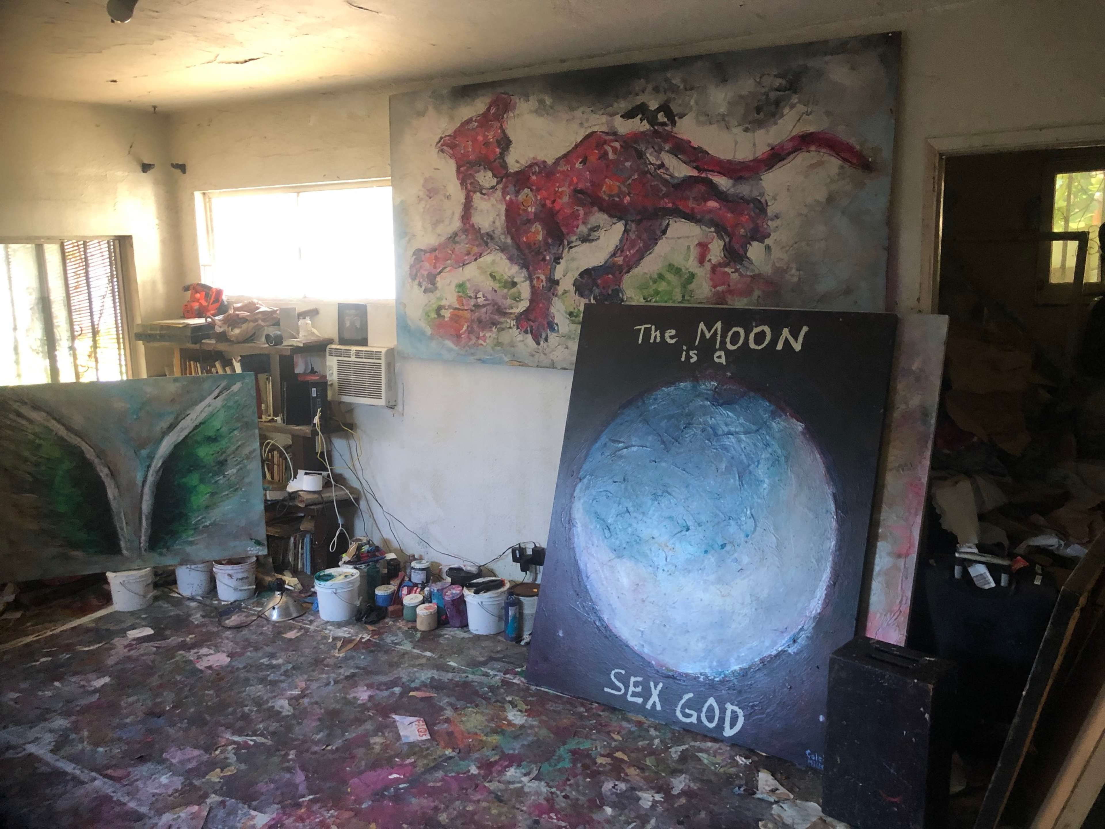 The image shows an artist's studio with colorful paintings, paint cans, and a cluttered floor.