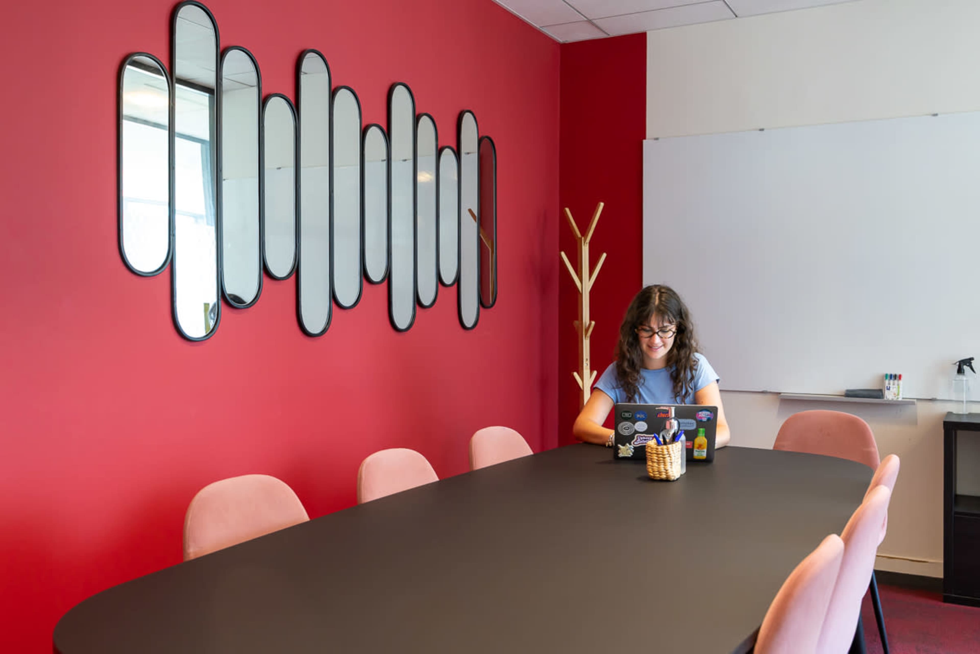 A woman is working on a laptop at a large black table in a conference room with a red wall and decorative mirrors.