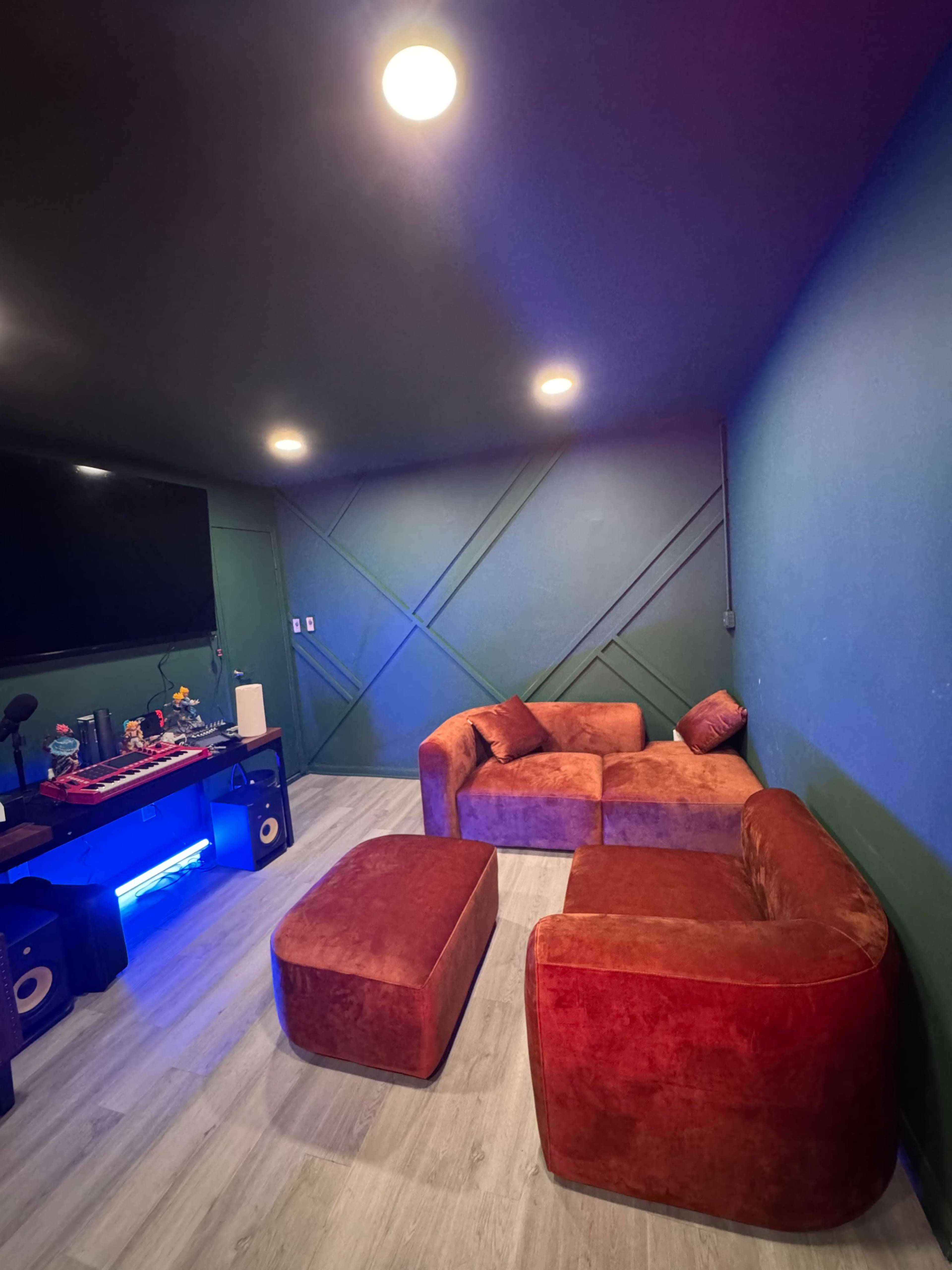 The image shows a small room with a green wall, featuring a red sectional sofa and an ottoman, along with a media setup on a black console.