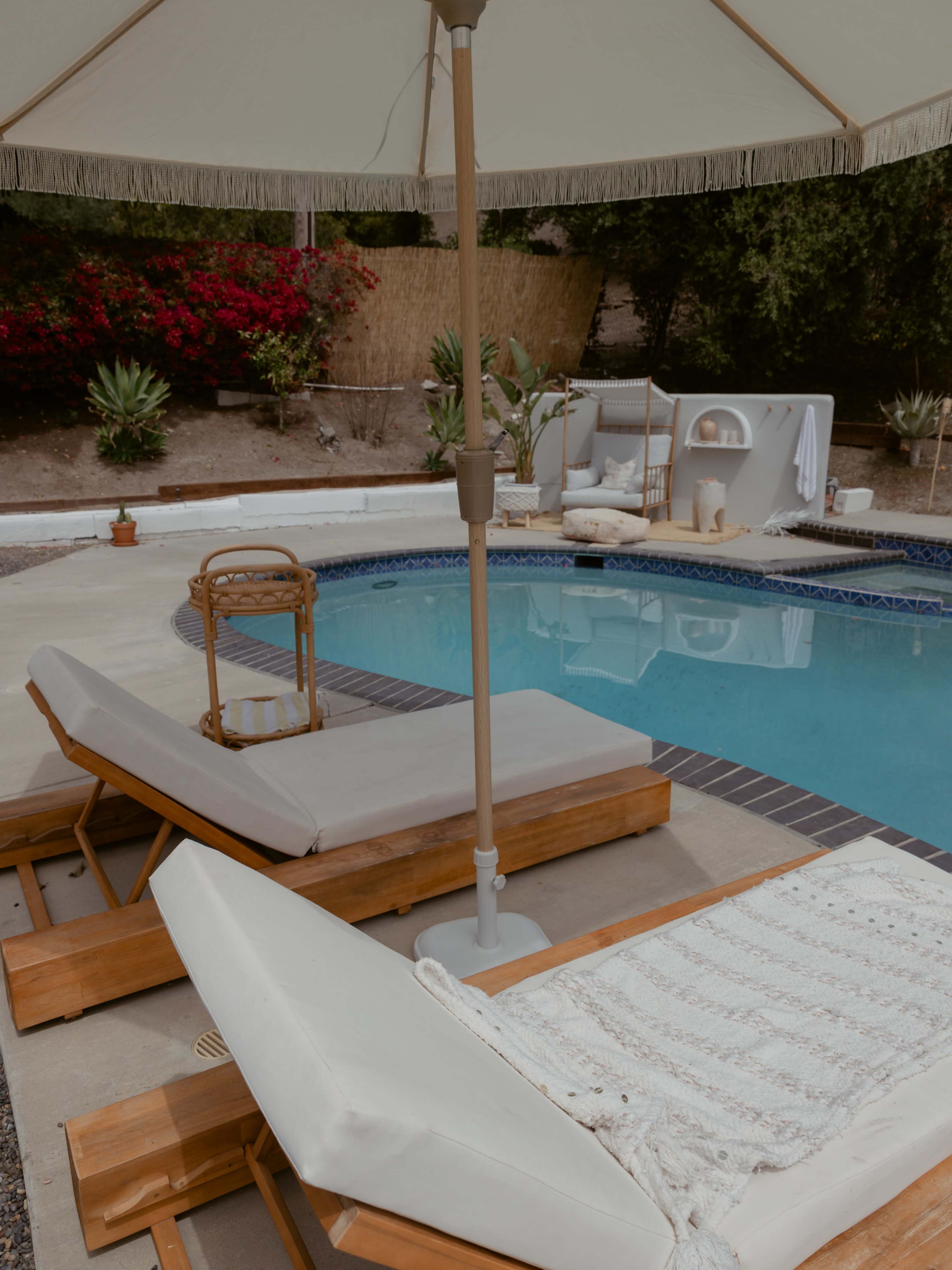Two beige lounge chairs on a pool deck, accompanied by an umbrella, beside a clear blue swimming pool surrounded by greenery and decorative plants.