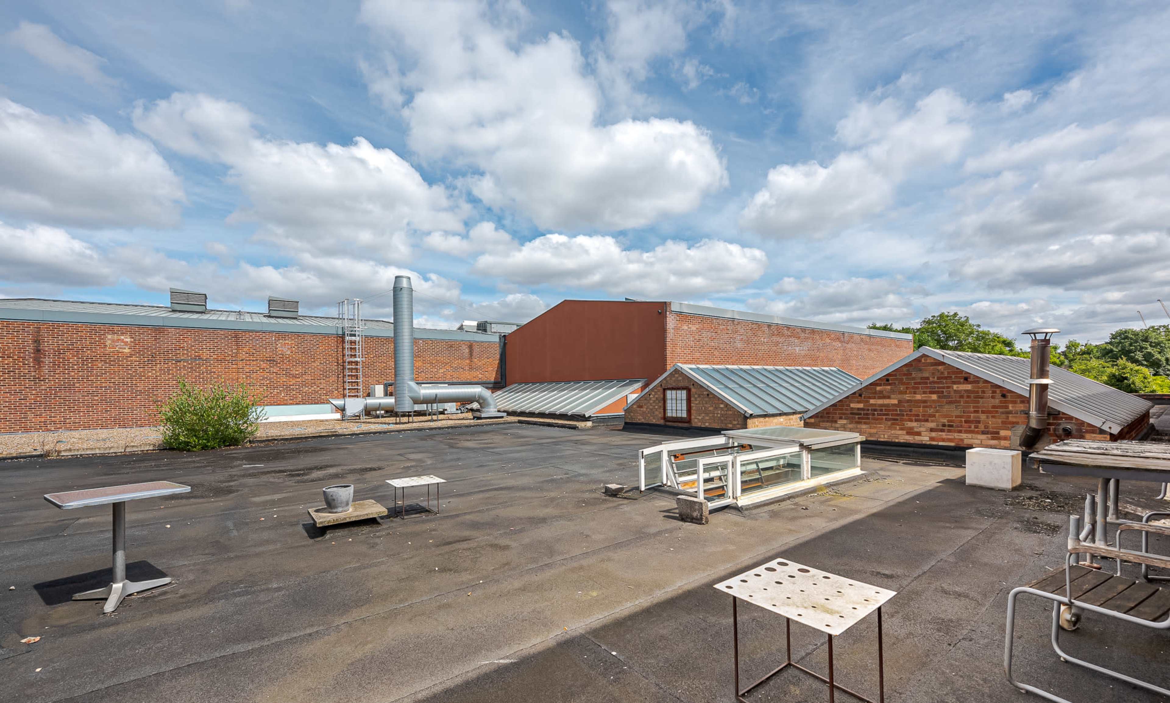 The image shows a flat rooftop with various structures, including ventilation pipes, green vegetation, and metal tables.