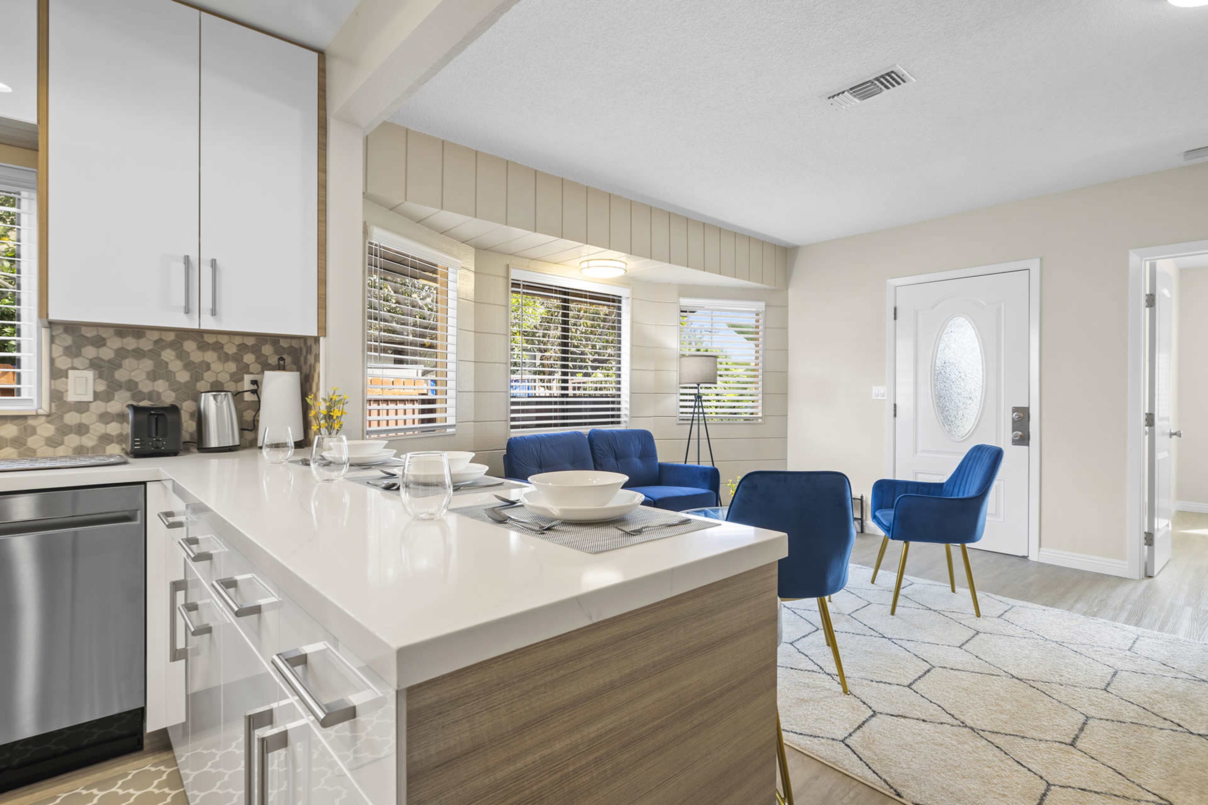 A modern kitchen with a dining area featuring a white countertop, stainless steel appliances, and a living room with blue chairs and large windows.