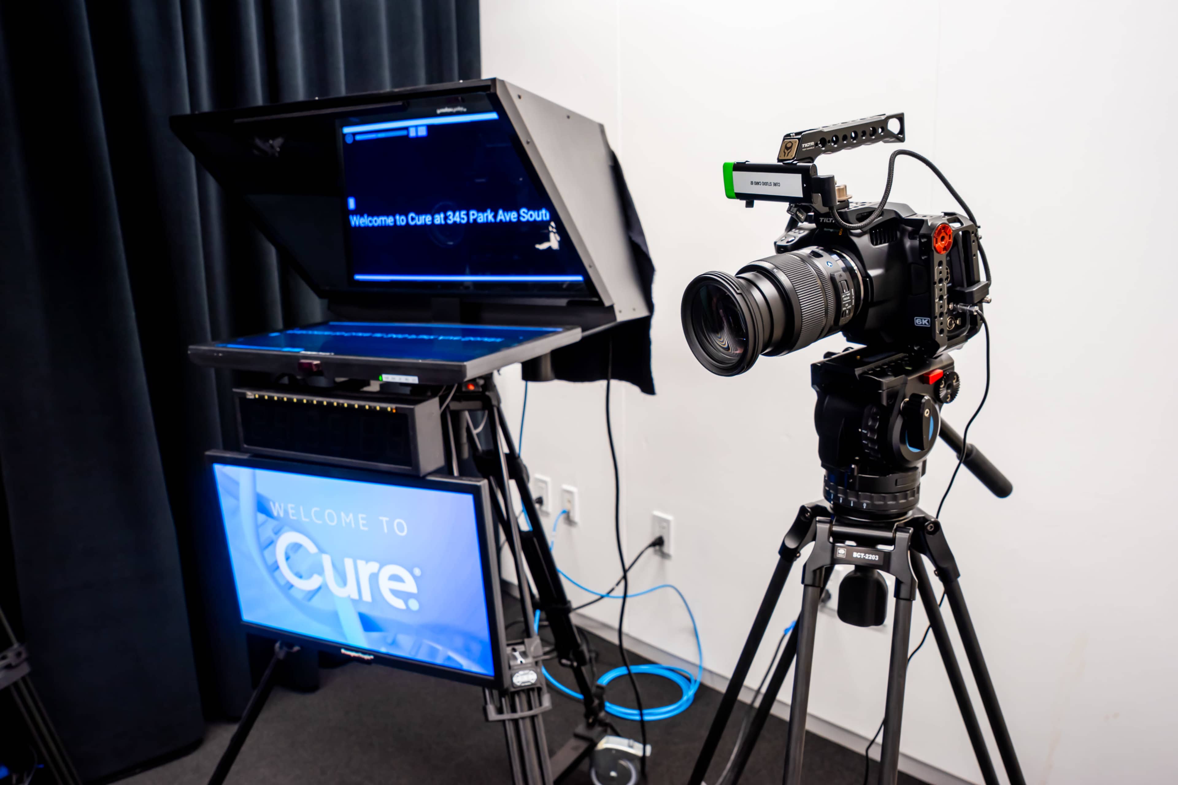 A camera mounted on a tripod stands next to a monitor displaying "Welcome to Cure" in a recording studio setup.