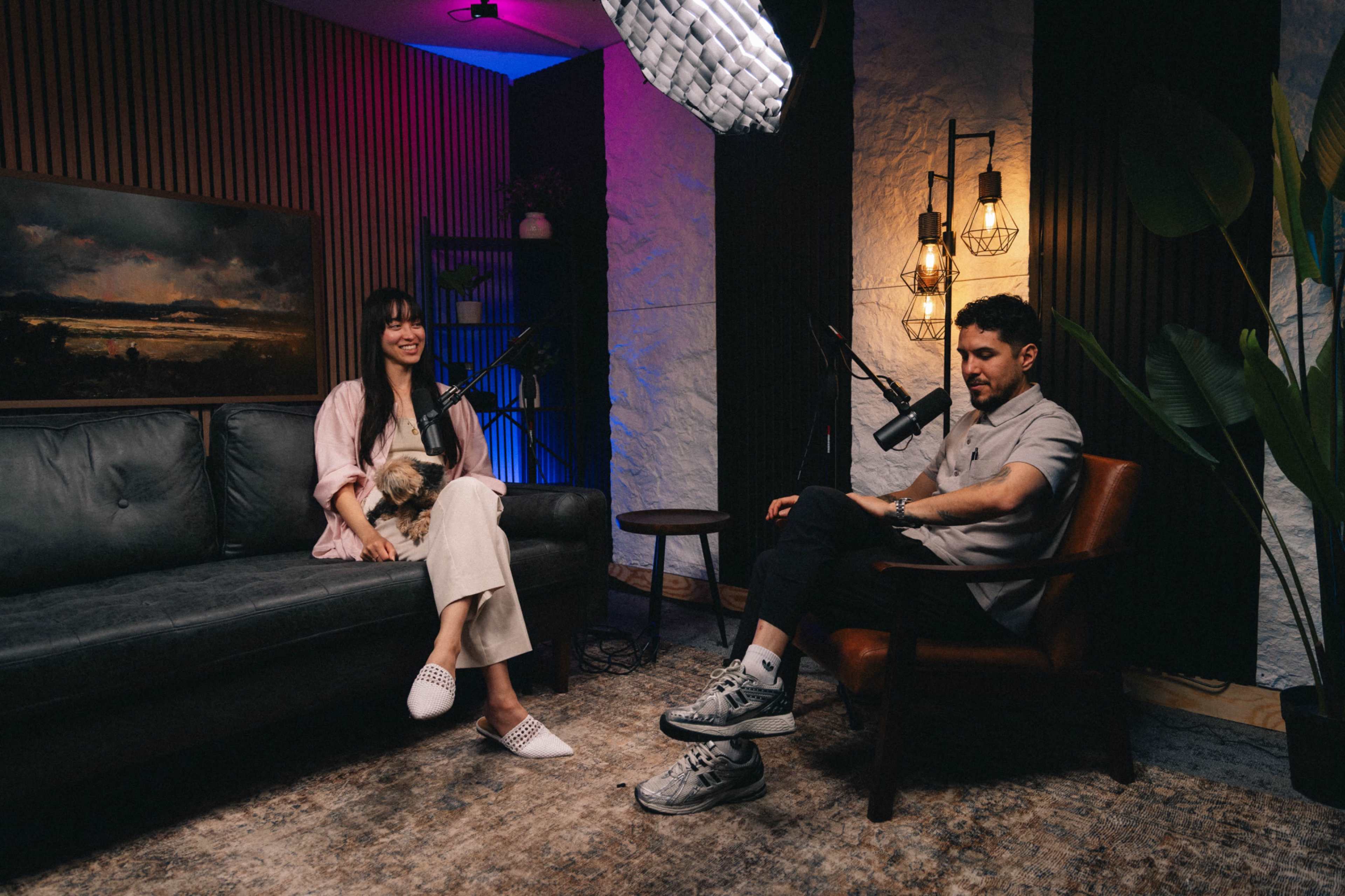 A woman with a small dog sits on a couch while a man sits across from her, both positioned in a well-lit room designed for a podcast recording.