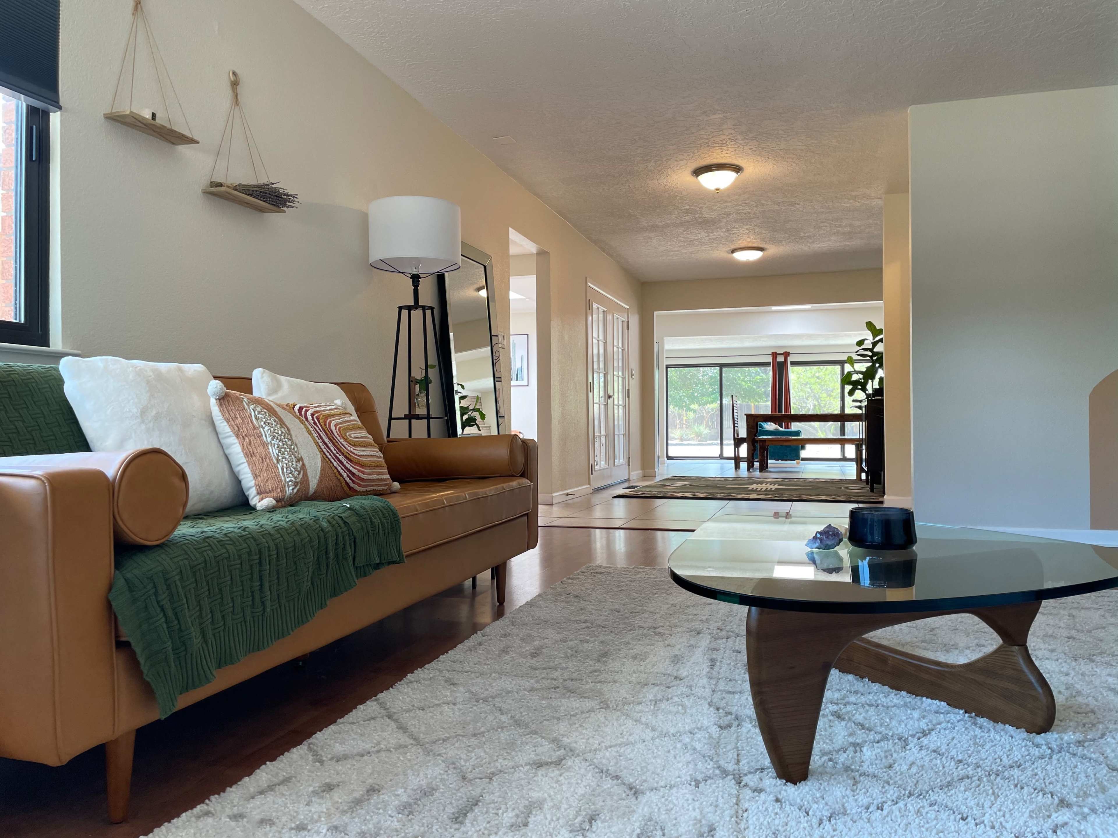A modern living room features a tan leather sofa with a green throw, a glass coffee table, and a view of an adjacent room with a dining table.