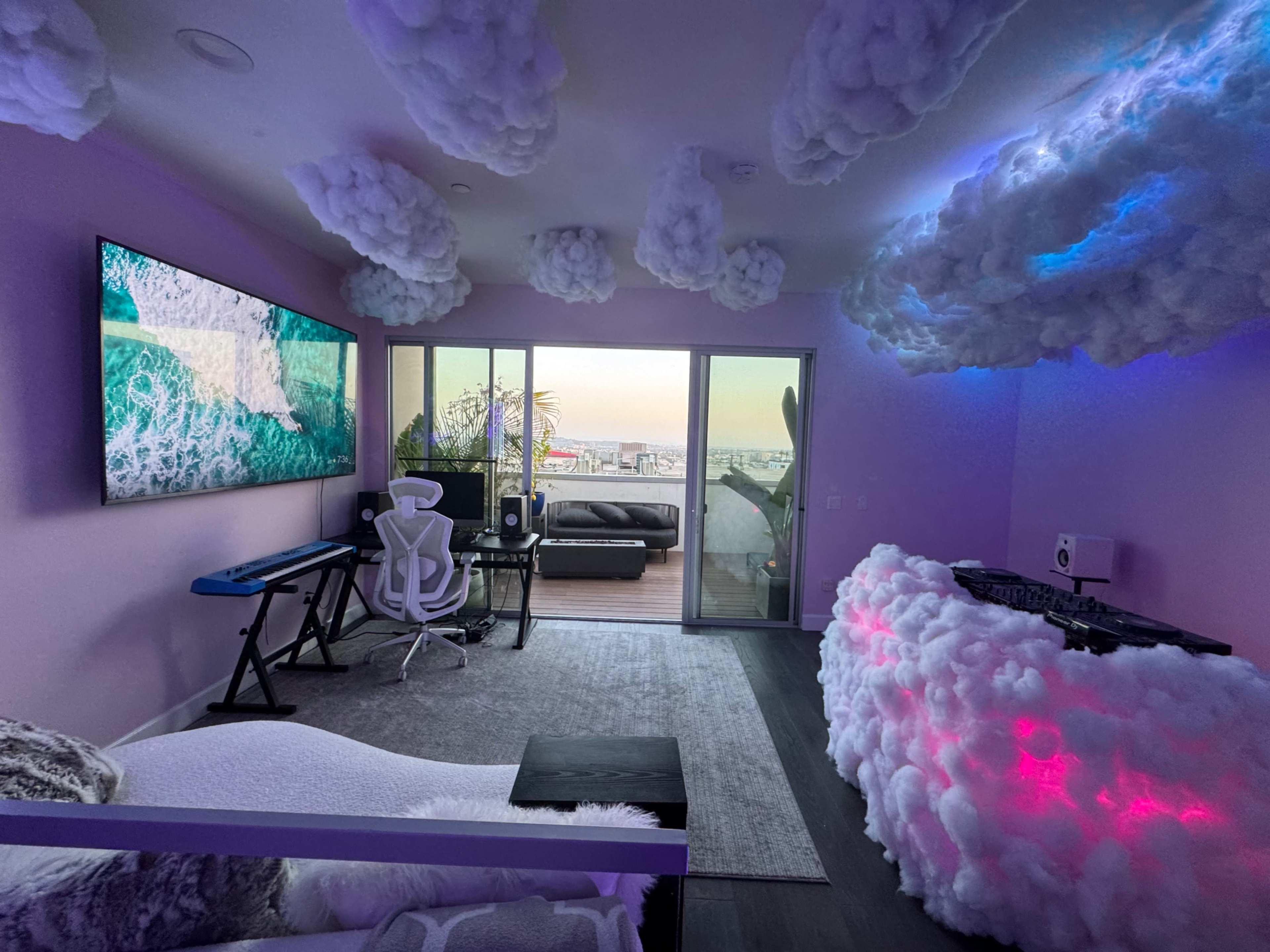 A modern room features a keyboard setup, a cloud-themed ceiling, a wall-mounted TV displaying ocean waves, and a balcony with a city view.