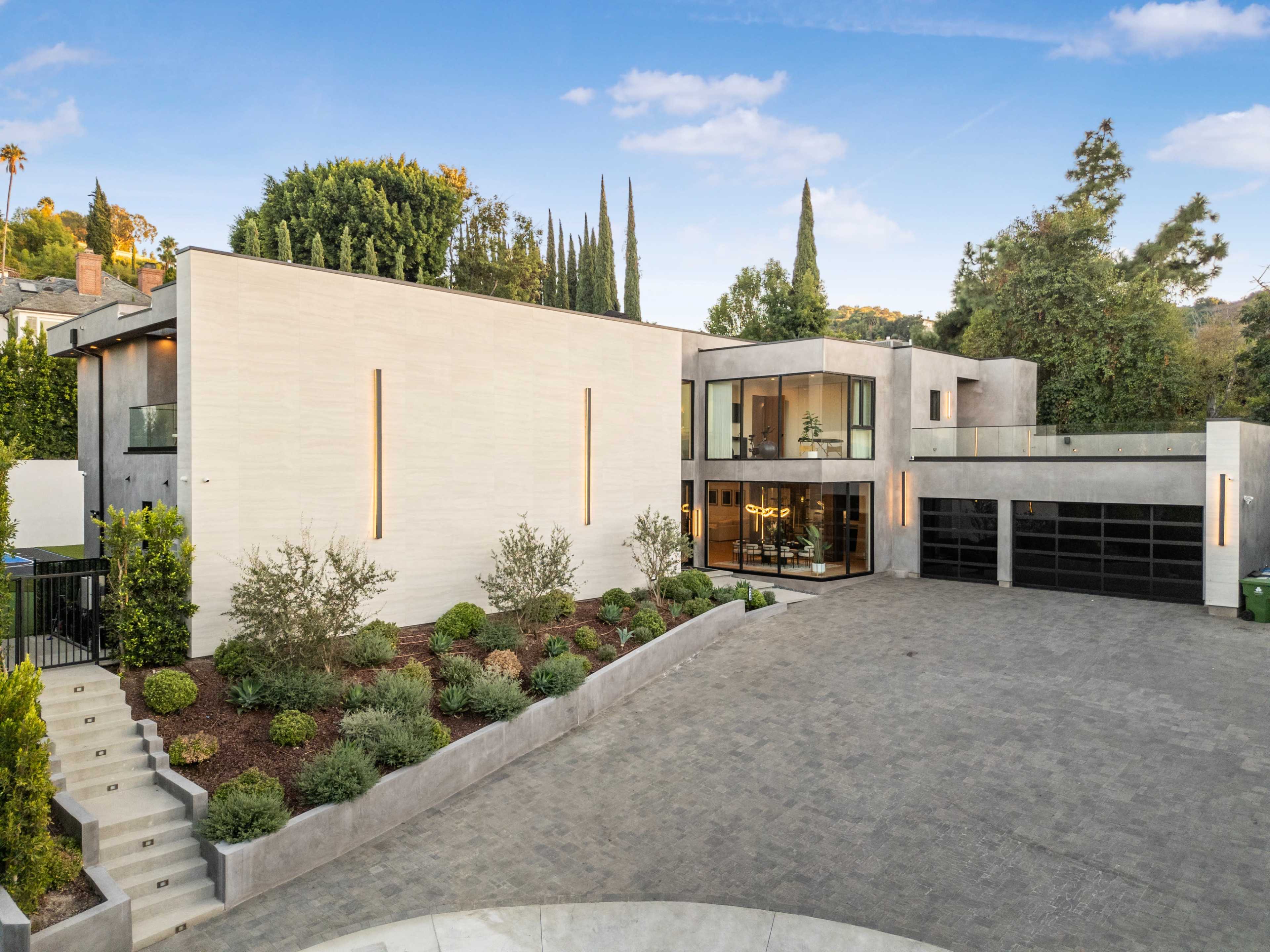 The image shows a modern, two-story house featuring large windows, a landscaped front yard, and a driveway made of cobblestones.