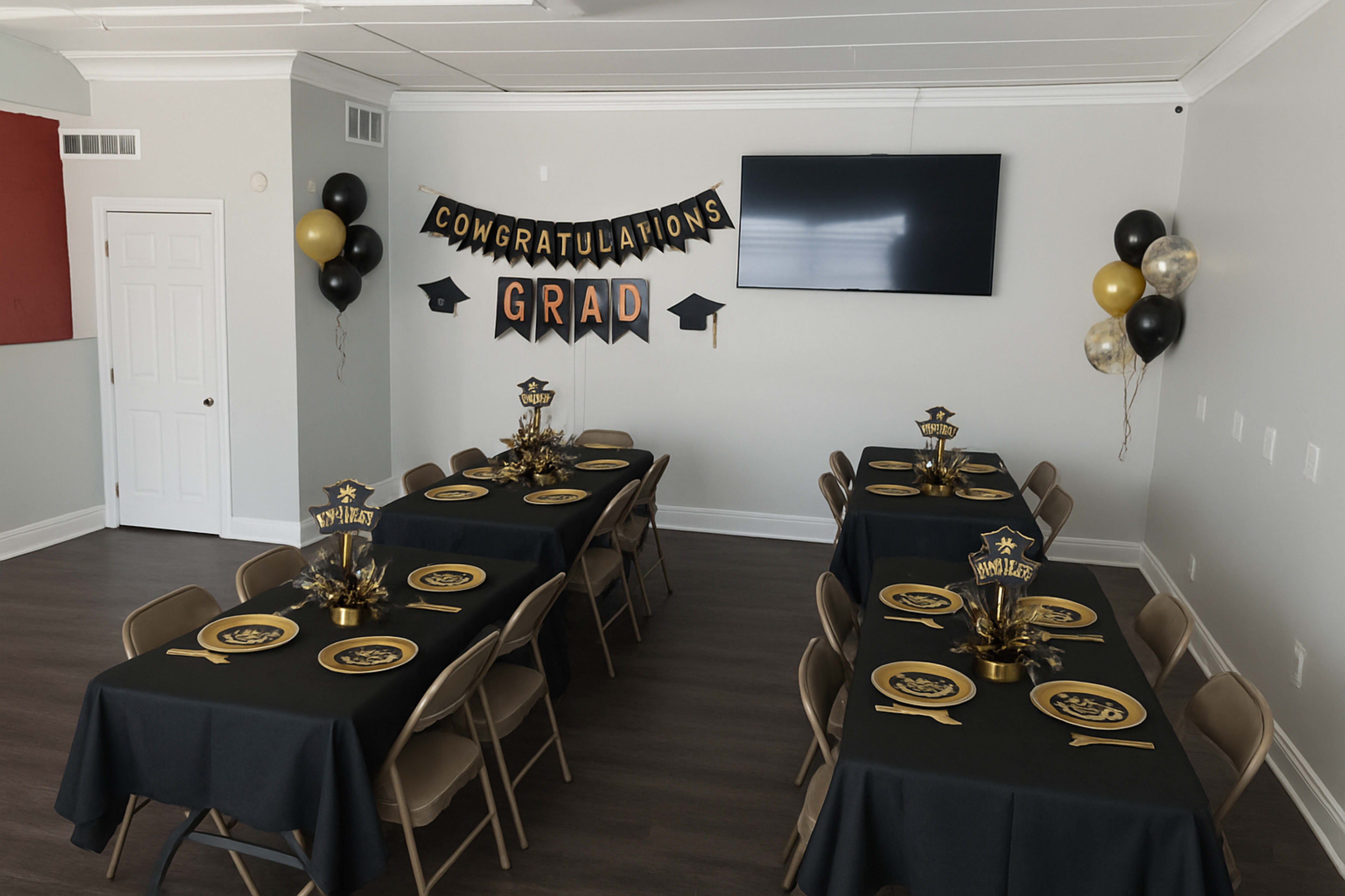 The image shows a decorated room with two long tables covered in black tablecloths, each set with gold plates and centerpieces, while a banner that reads "CONGRATULATIONS GRAD" hangs on the wall above a television.