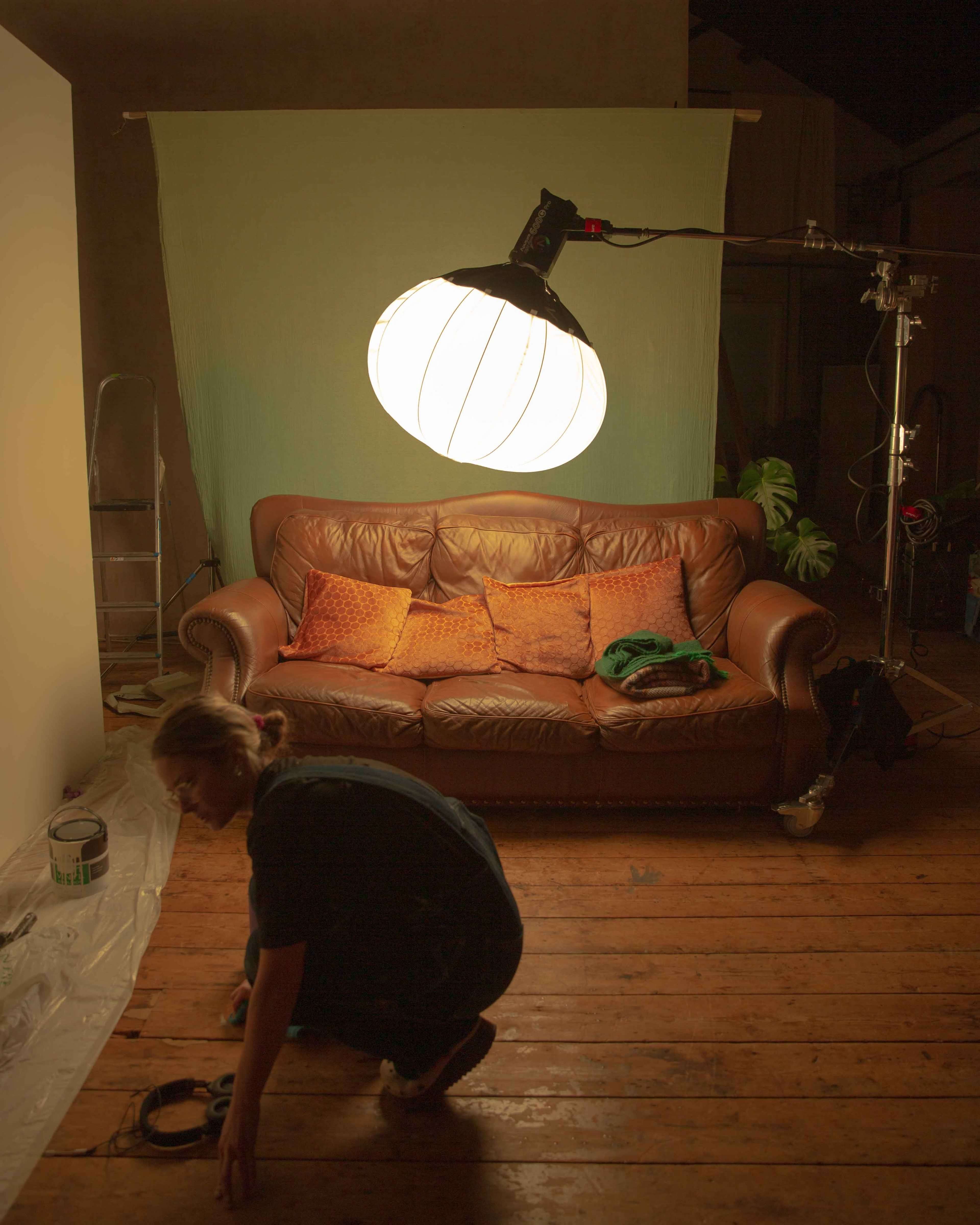 A person is crouched on the wooden floor near a leather sofa with decorative cushions, while an overhead light illuminates the scene.