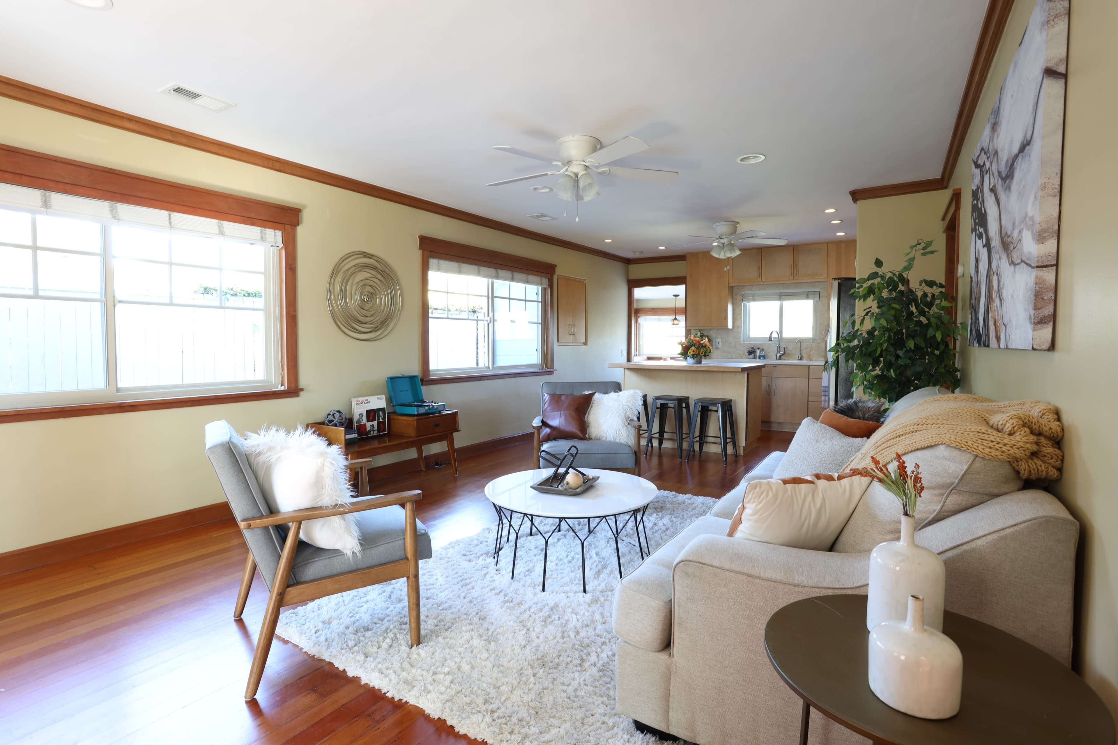 A light-filled living room features a cozy sectional sofa, a round coffee table, and a textured area rug, with a kitchen in the background.