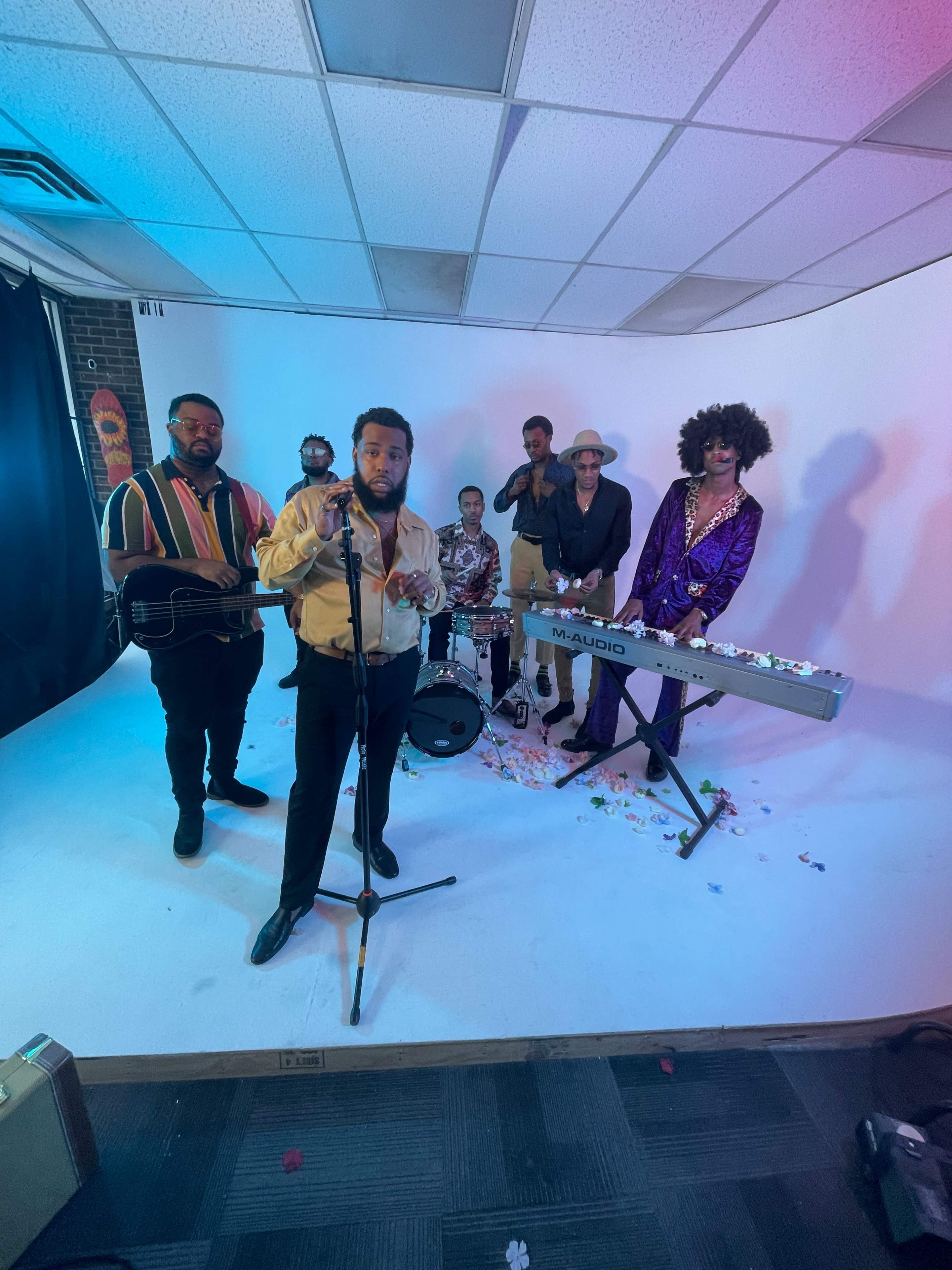 A seven-member band performs on a small stage with a white backdrop, featuring instruments like a keyboard and drums, while scattered flower petals cover the floor.