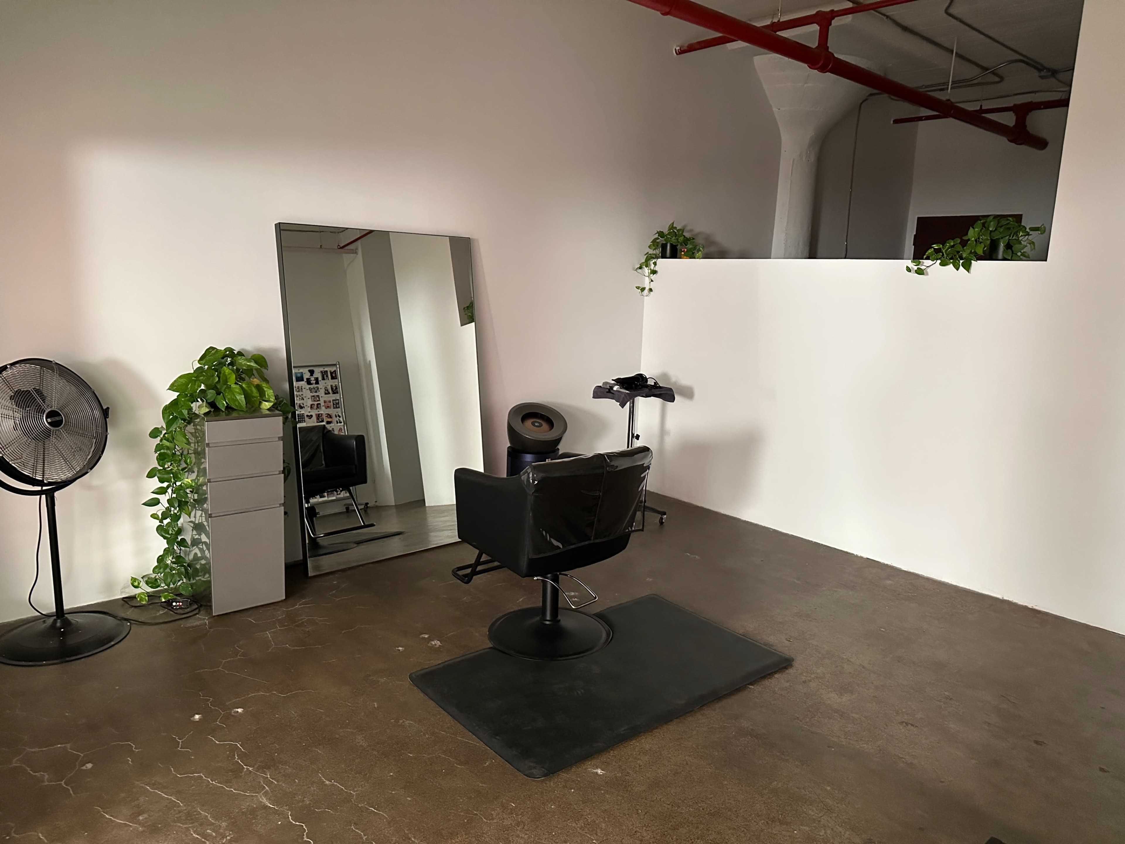 The image shows a minimalist hair salon interior featuring a black salon chair, a large mirror, a potted plant, and a fan in a spacious room with concrete flooring.