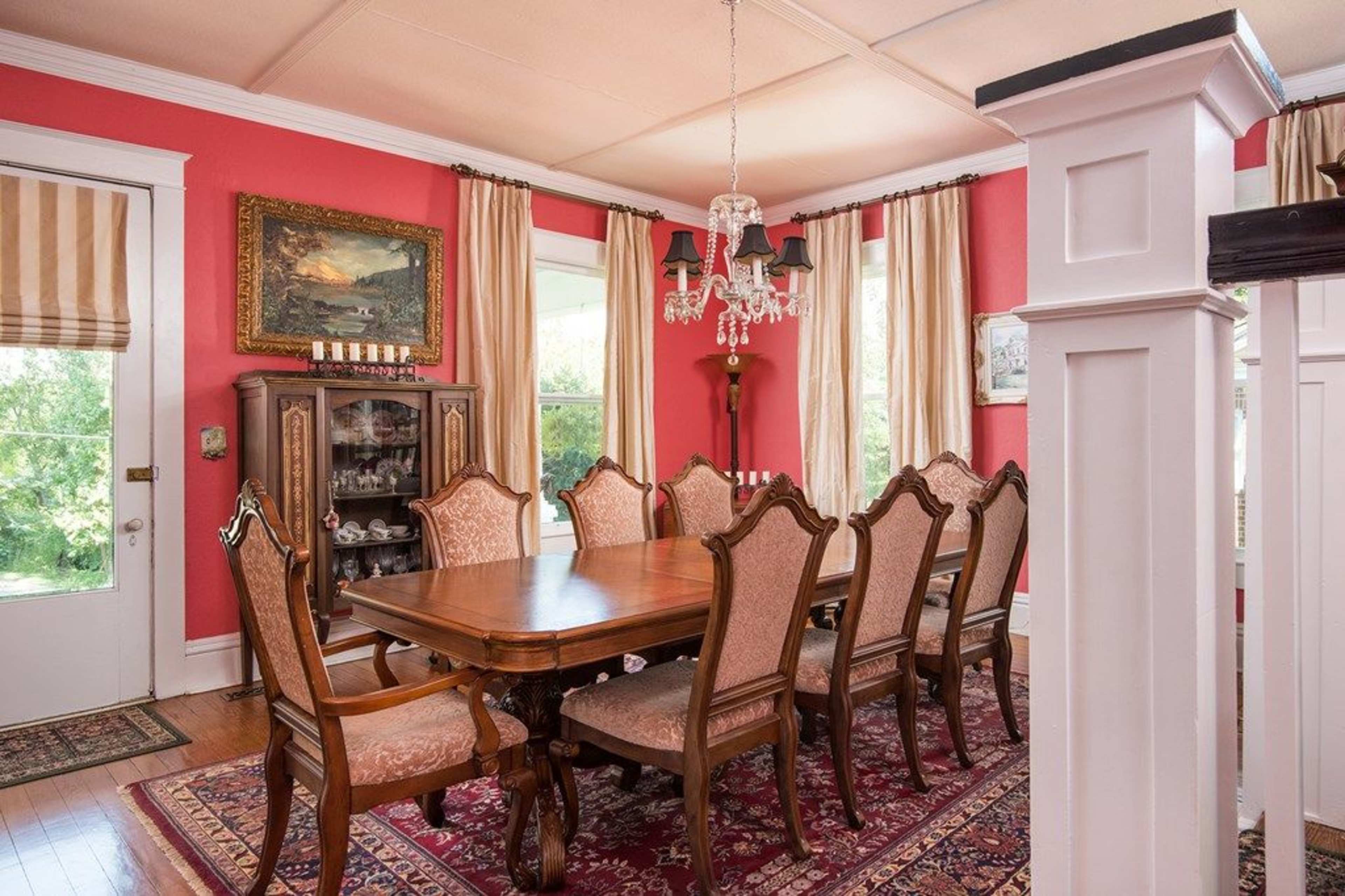A dining room with a wooden table surrounded by eight upholstered chairs, bright red walls, and a chandelier hanging from the ceiling.
